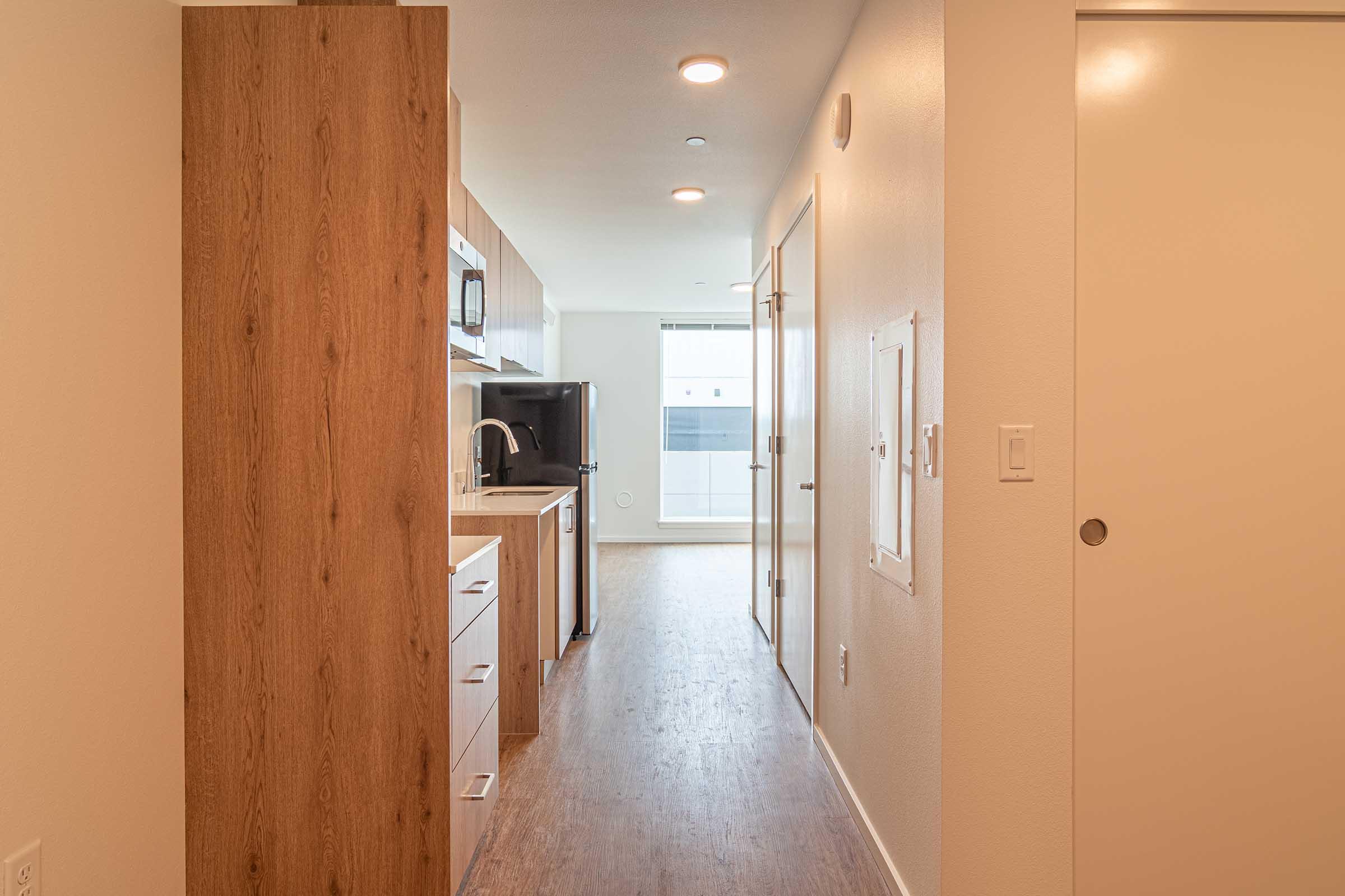 A modern hallway in an apartment, featuring wooden cabinets on one side and a kitchenette with a microwave and sink. The floor has light wood finish, and there are doors leading to other areas. Bright lighting enhances the spacious feel, and a large window at the end opens to an outside view.