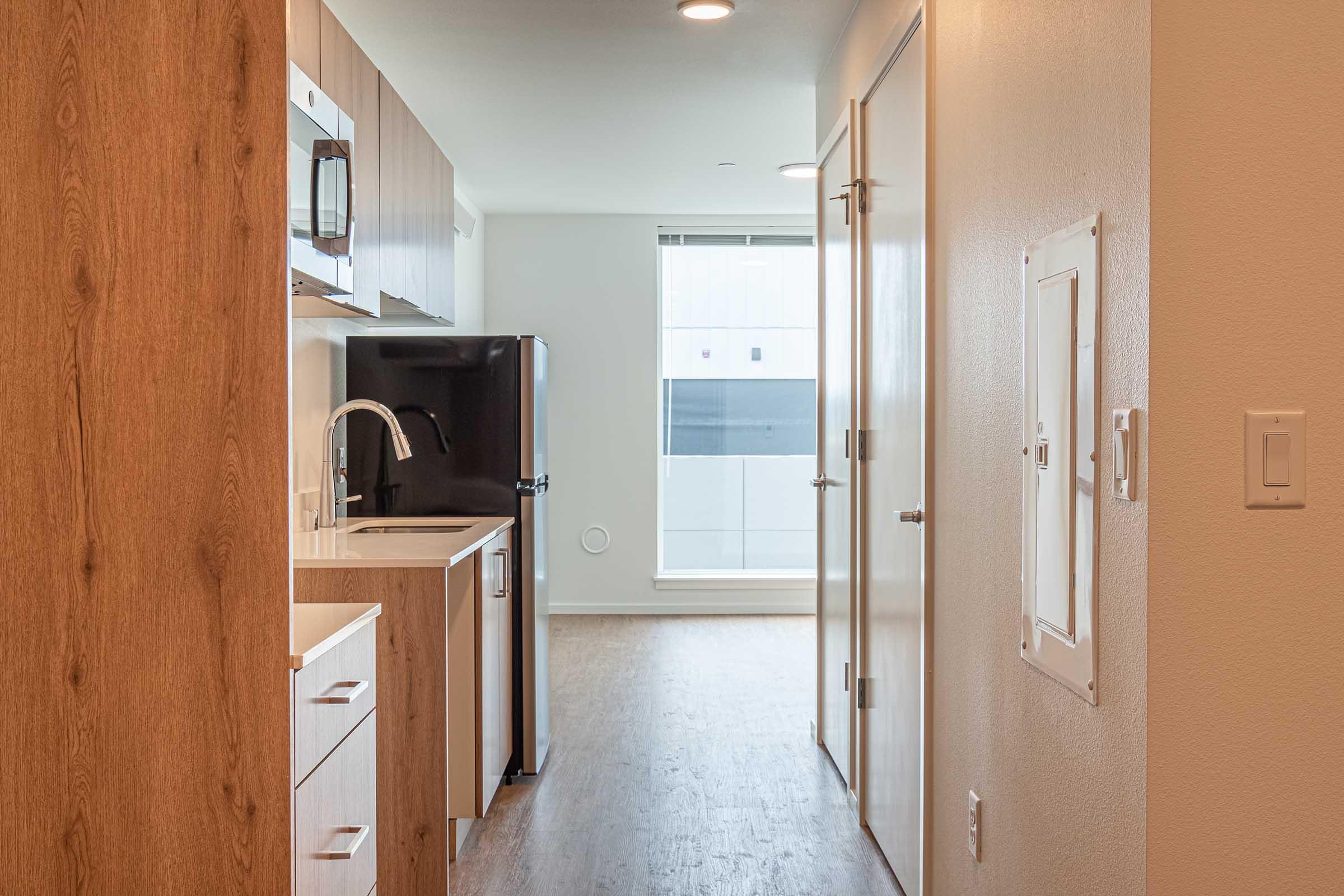 A modern kitchen area with wood cabinetry and stainless steel appliances. A hallway leads to a brightly lit room with large windows. An open floor plan allows for easy movement, with a refrigerator, microwave, and sink visible. Light hardwood flooring enhances the contemporary feel of the space.