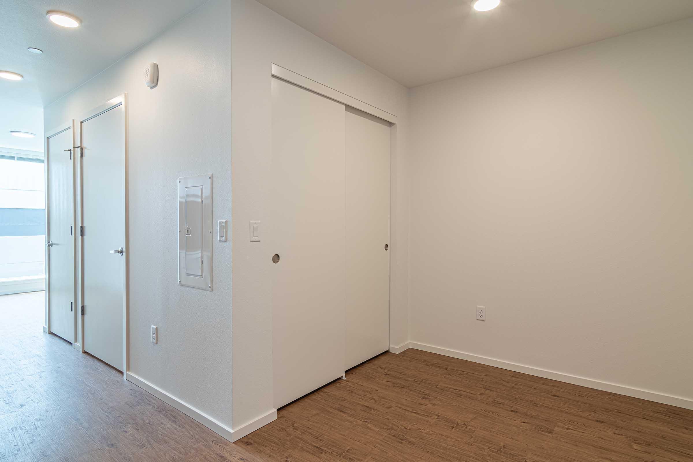 A clean and modern interior hallway featuring white walls, a wooden floor, and two closed doors. The left door is in a nook with an electrical panel next to it, while the right door is a sliding closet door. Soft lighting from recessed fixtures enhances the bright atmosphere.