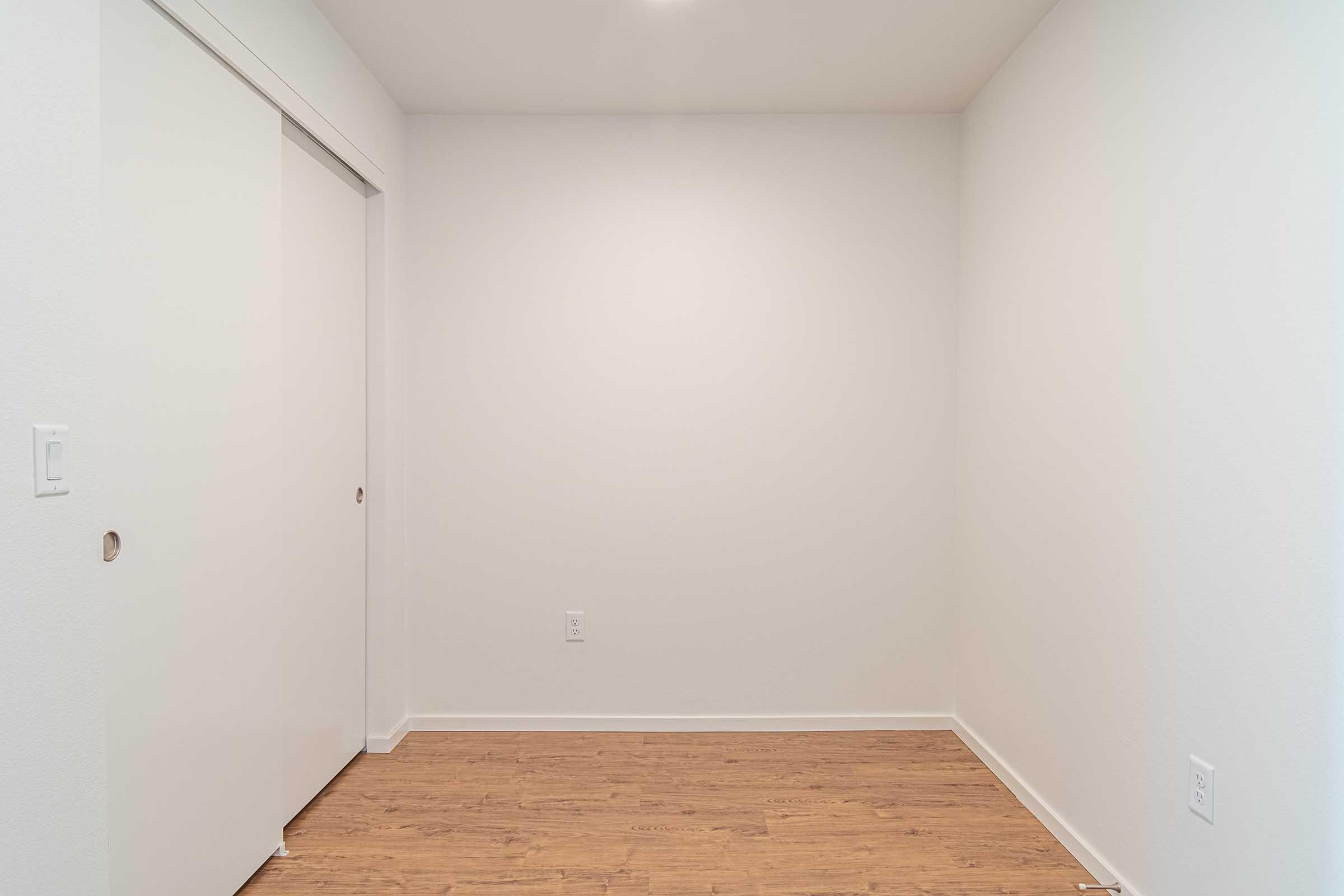 A minimalistic empty room with light-colored walls and a wooden floor. A closet door is visible on the left side, and the space is well-lit but devoid of furniture or decoration, creating a clean and open atmosphere.