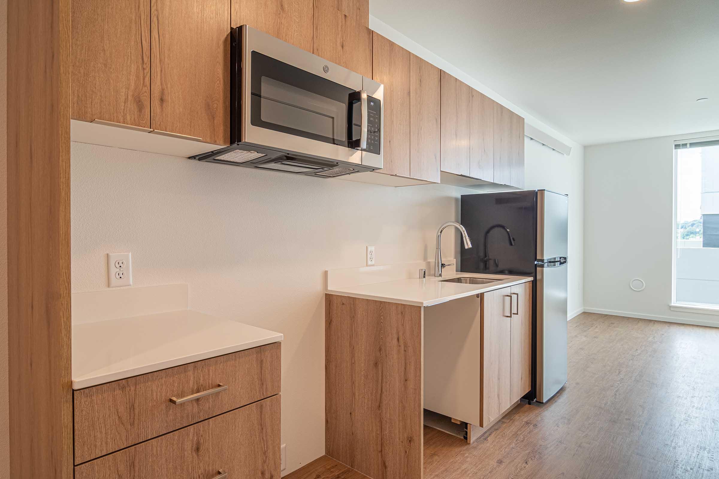 Modern kitchen with wood-paneled cabinetry, a stainless steel microwave above a white countertop, a sink, and a refrigerator. The space features an open layout with natural light coming from a large window, showcasing a clean and minimalist design.