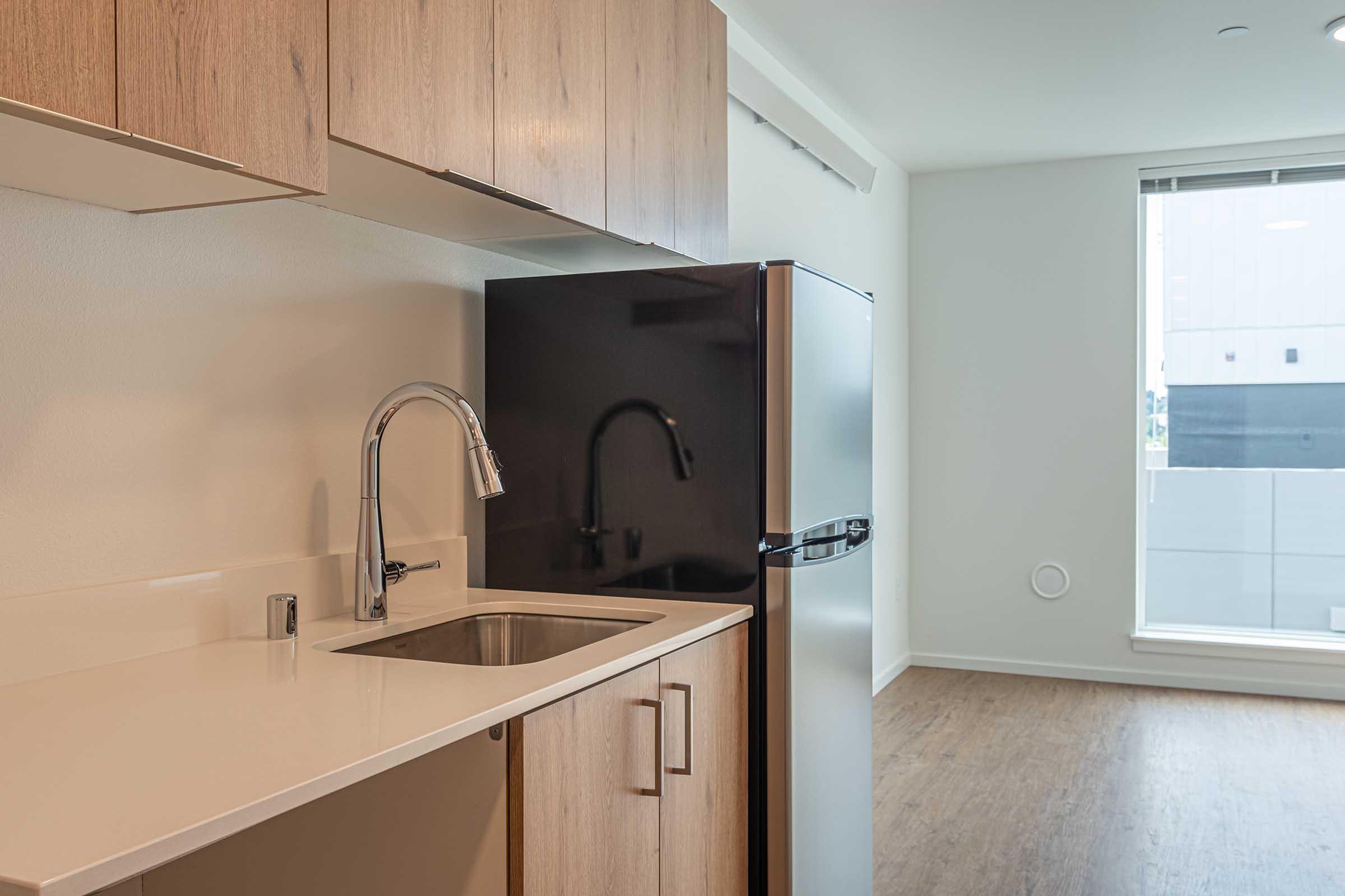 A modern kitchen featuring a stainless steel sink with a sleek faucet, a black refrigerator, and wood cabinetry. The walls are painted in a light color, and the flooring is a warm wood tone. Natural light enters through a window, illuminating the minimalist design and open space.