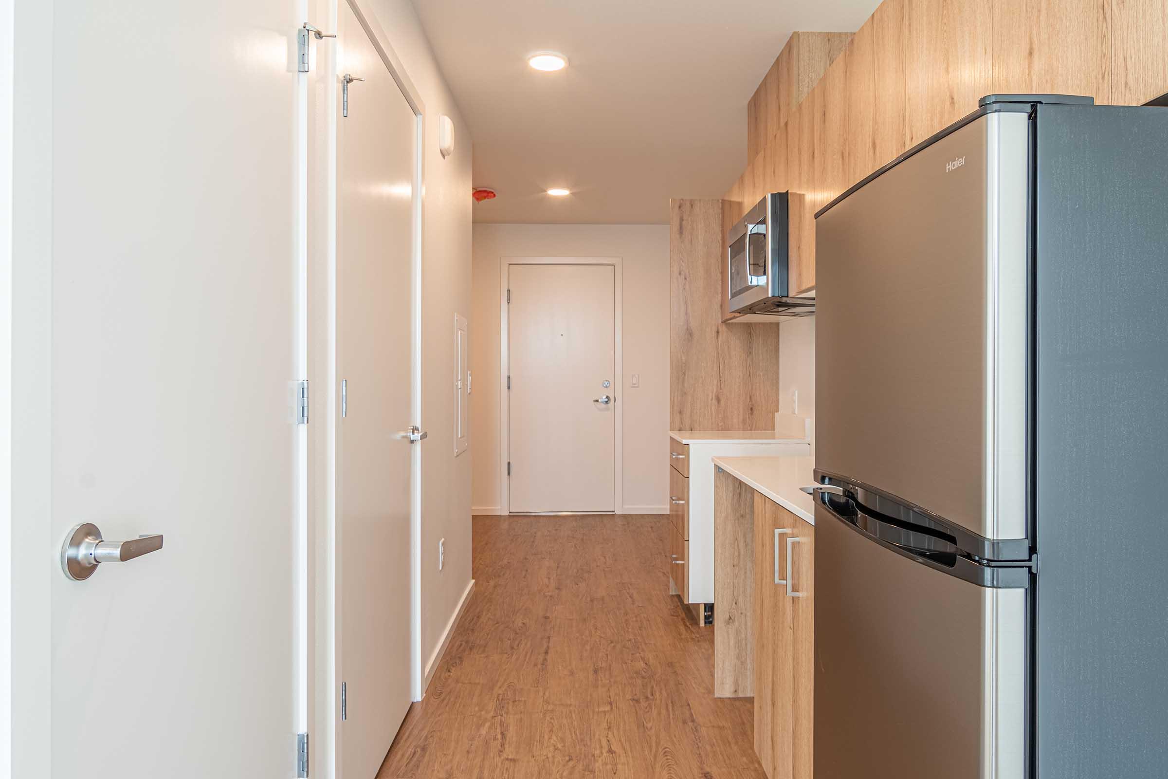 A modern kitchen area featuring a stainless steel refrigerator, a built-in microwave, and wooden cabinetry. The hallway leads to a door, with light-colored walls and a wooden floor, creating a spacious and inviting atmosphere.