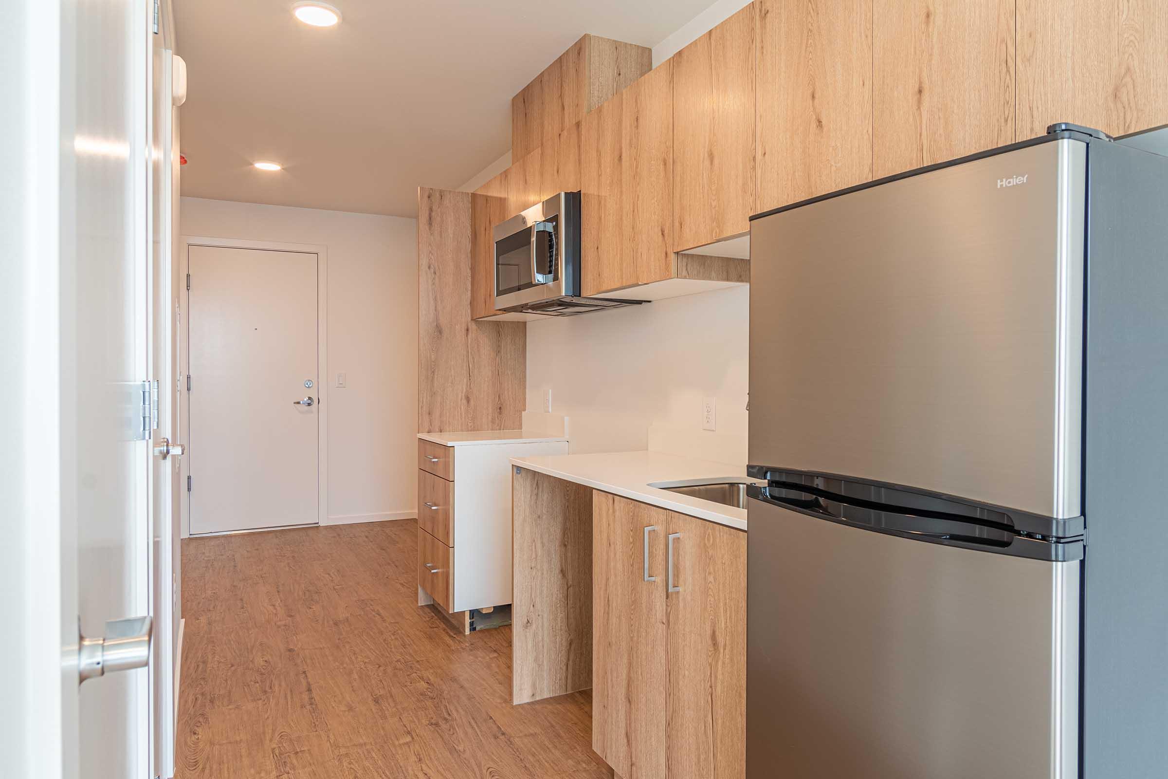 A modern kitchen featuring wooden cabinetry, a stainless steel refrigerator, a microwave above the stove, and a minimalist countertop. The space has an open layout with a view of the entrance and light wood flooring, creating a bright and inviting atmosphere.