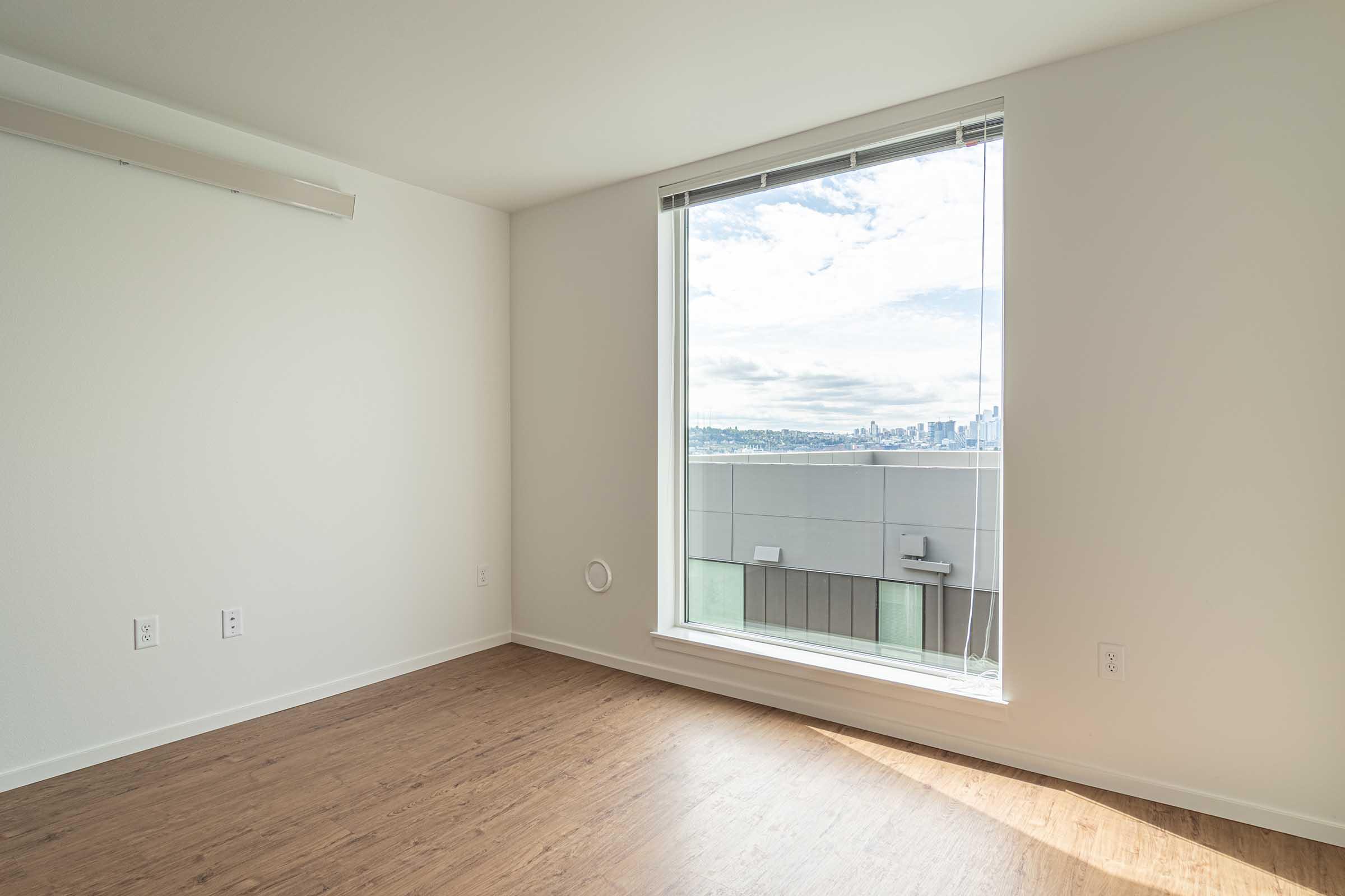 Empty room with large window offering a view of a city skyline. The walls are light-colored, and the flooring is wood-like. The space is bright and minimalist, featuring a small circular vent on one wall. Natural light fills the room, highlighting the clean lines and open feel.