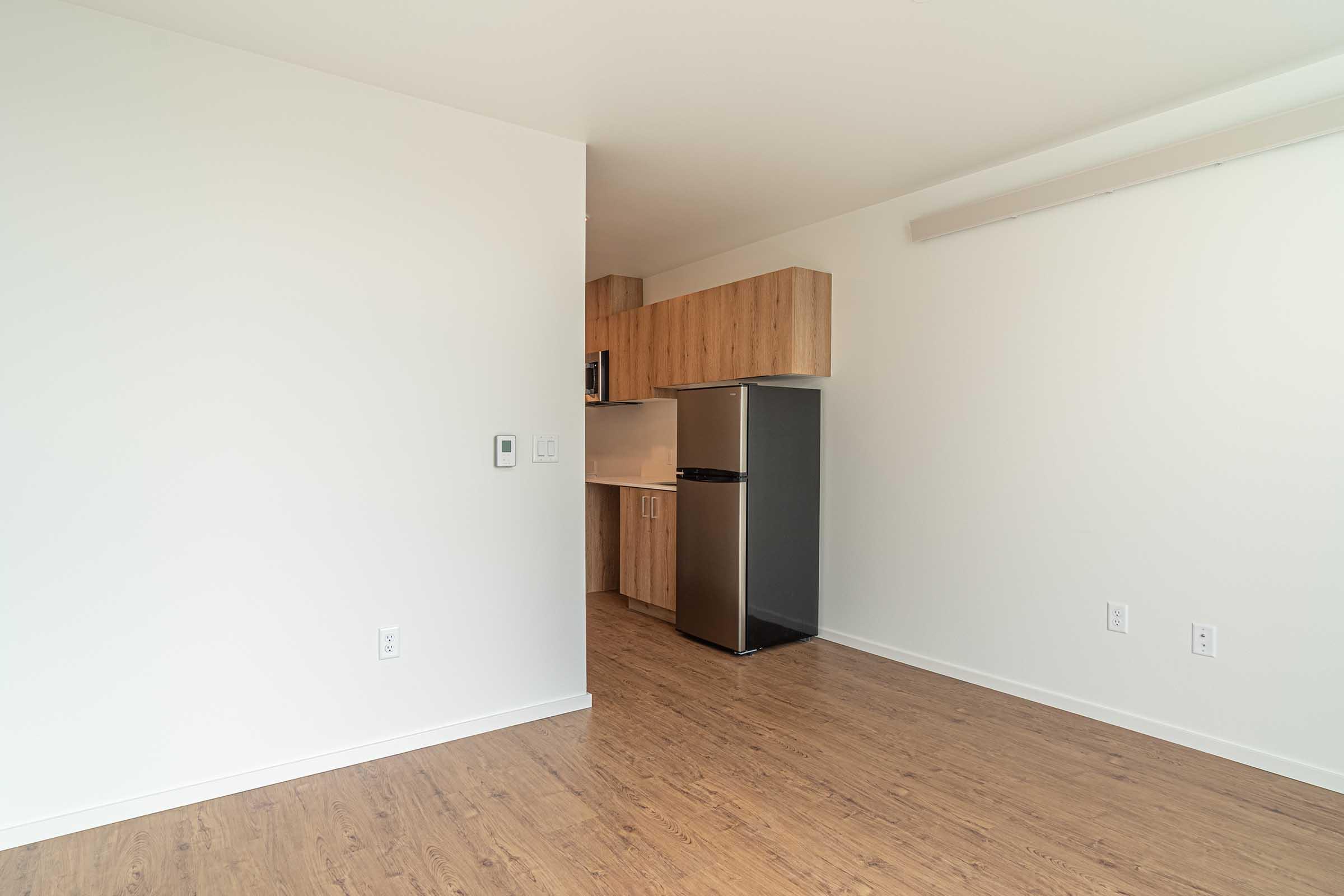 Modern studio apartment with light wood flooring, featuring a kitchenette with wooden cabinets and a silver fridge. The walls are painted white, providing a bright and open atmosphere. The space is sparsely furnished, creating a minimalist aesthetic.