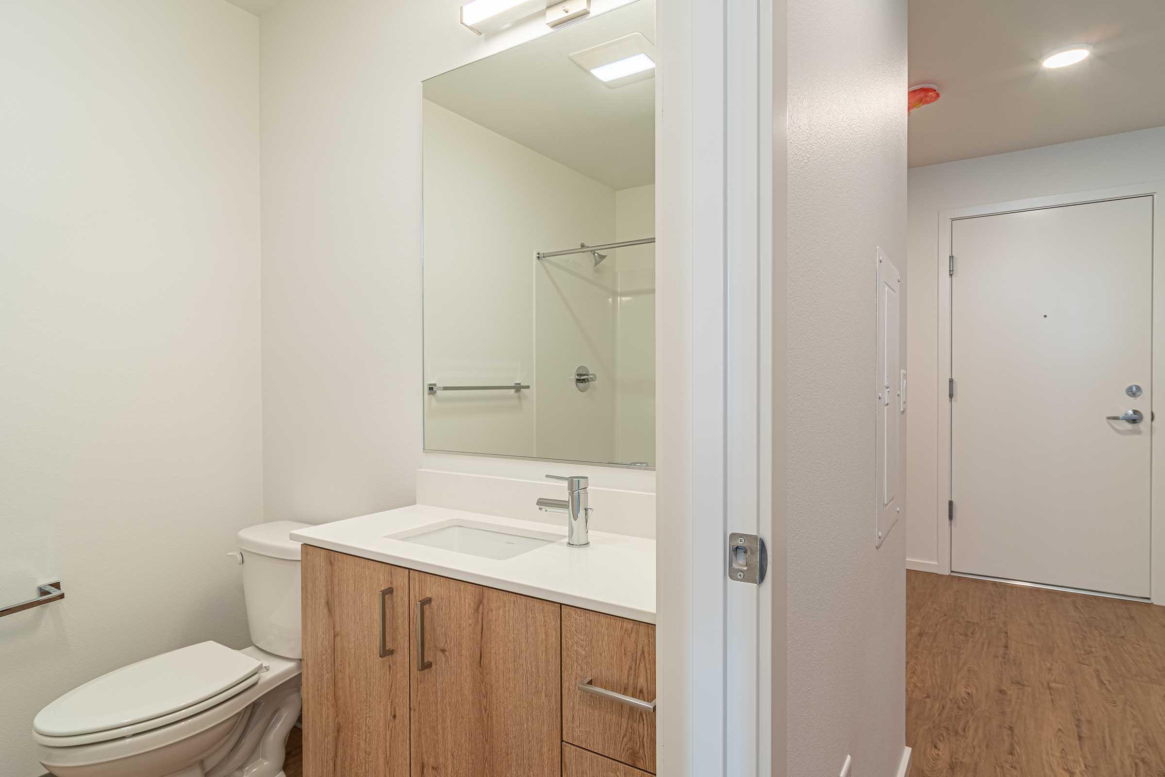 A modern bathroom featuring a toilet, a vanity with a sink and wooden cabinetry, a large mirror above the sink, and a glass shower enclosure. The walls are painted bright and the flooring is a light wood. A door leads to another room, with a fire alarm visible on the ceiling.