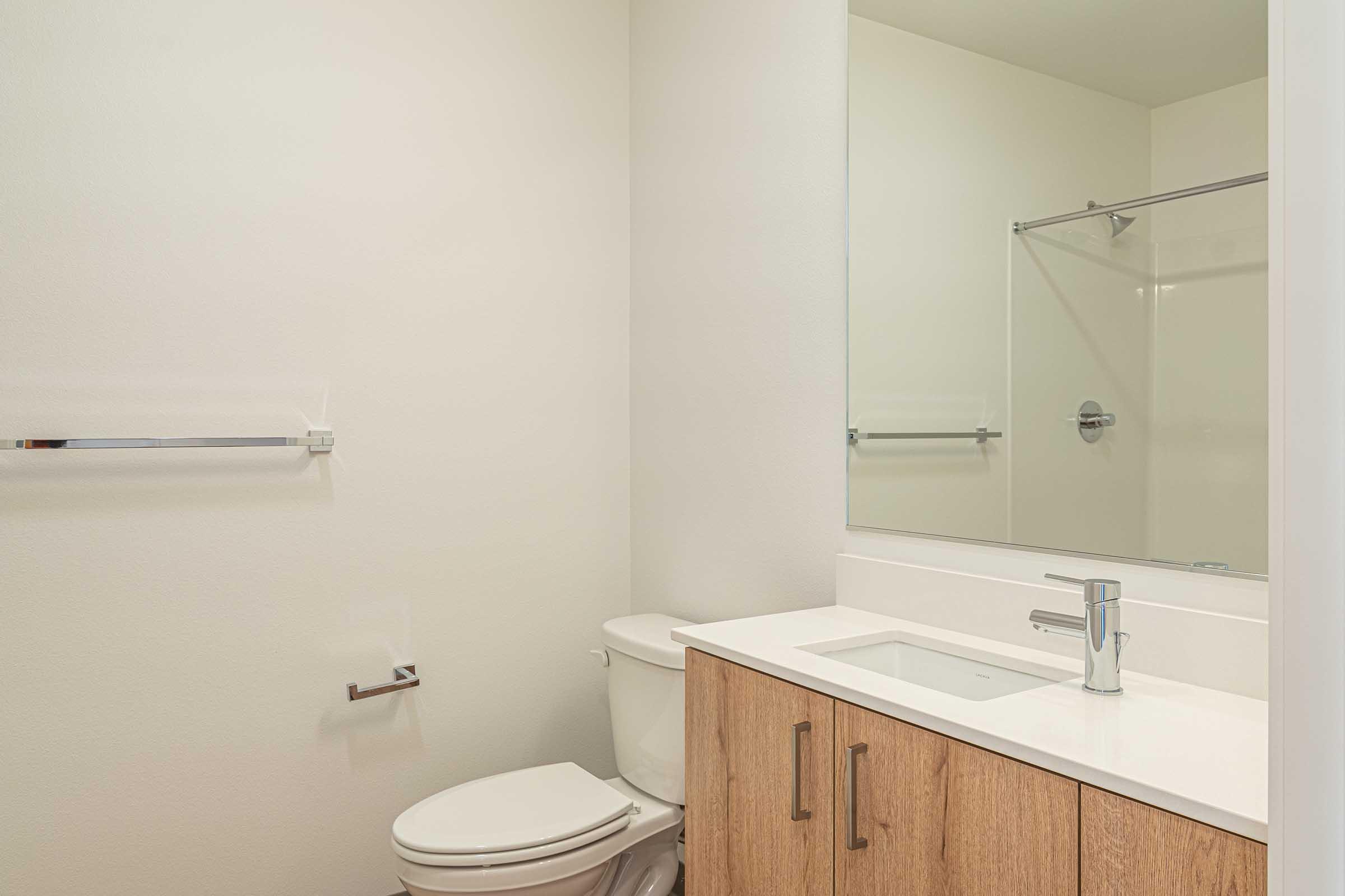 A modern bathroom featuring a white wall, a glass shower enclosure, a wooden vanity with a sink, and a toilet. A mirror hangs above the vanity, and a towel bar is mounted on the wall. The design is minimalistic and clean, with neutral colors and simple fixtures.