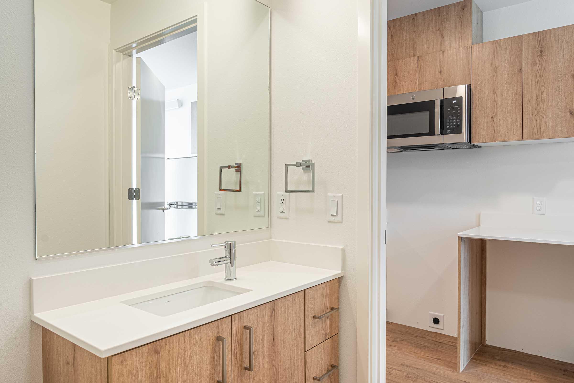 Modern bathroom with a clean white countertop, a sleek faucet, and wooden cabinetry. A large mirror hangs above the sink. In the background, a kitchenette area is visible, featuring a microwave and a small counter space, set against light-colored walls and wooden accents.