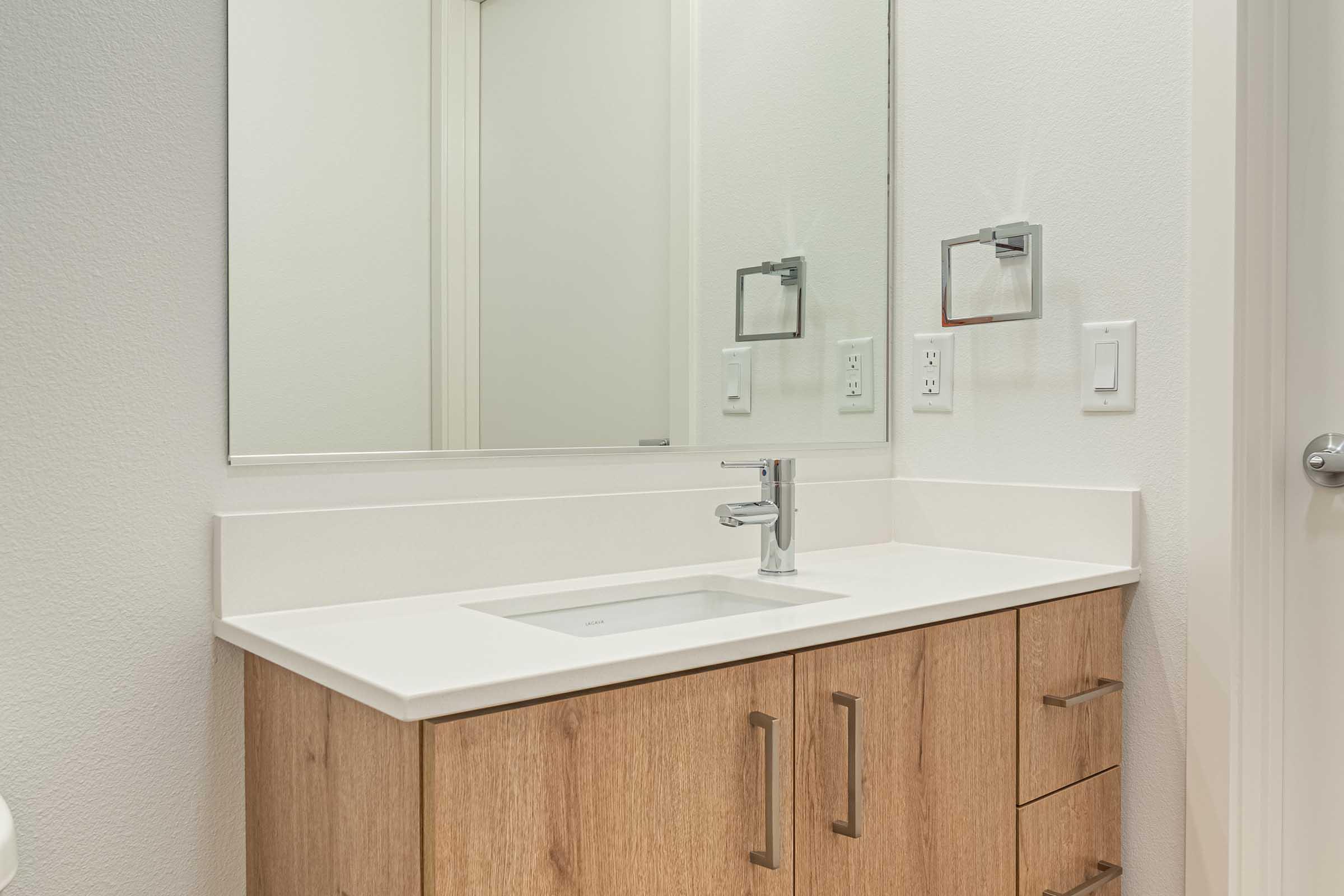 A modern bathroom featuring a clean, bright design. The countertop is white with a rectangular sink, complemented by a sleek faucet. There are wooden cabinet drawers below, and a large mirror above the sink reflecting the bright space. The walls are a neutral color, enhancing the contemporary feel.