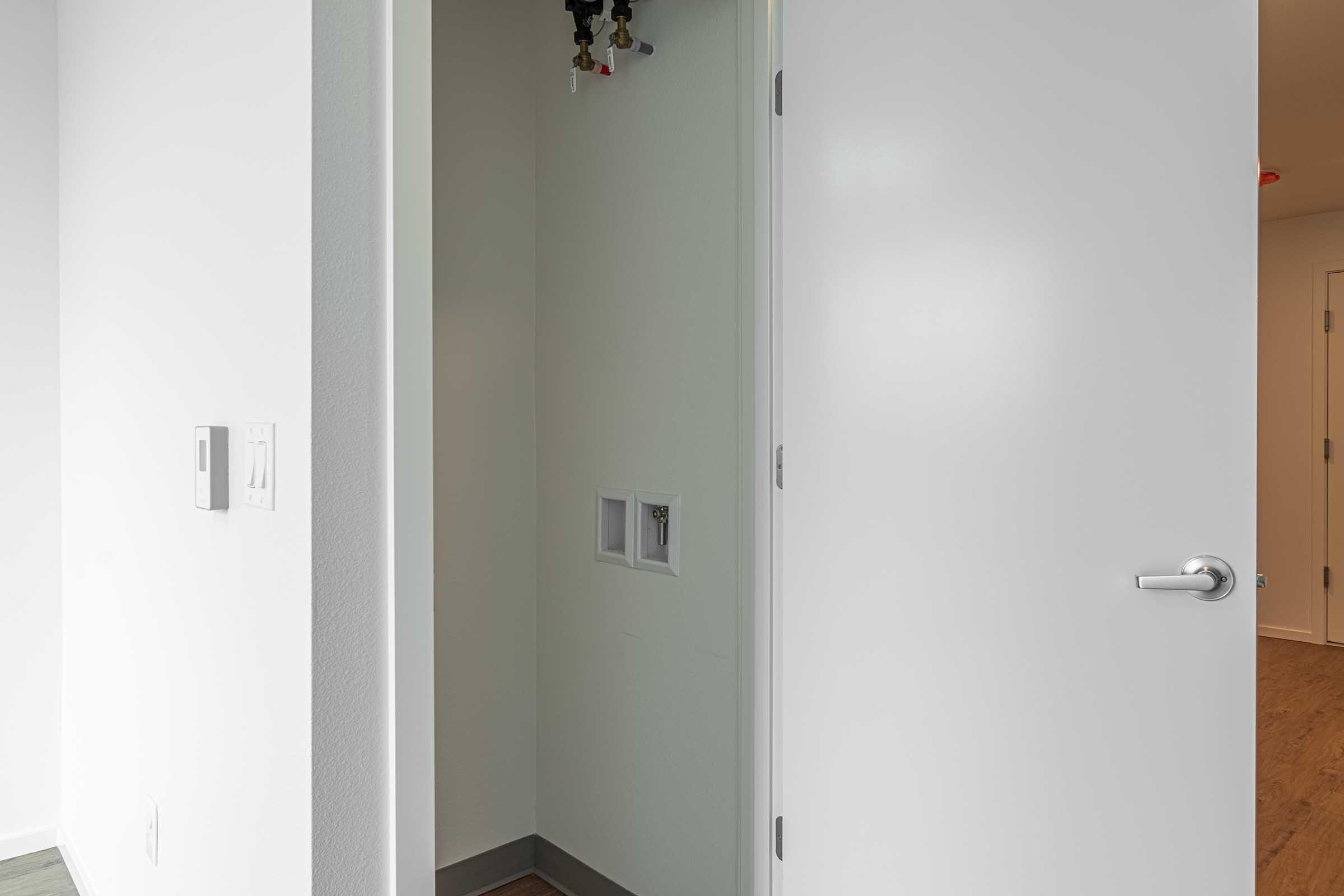 An empty corner of a room with a light-colored wall and two door openings. One door is partially open, revealing a narrow, unadorned space. It has plumbing connections visible in the wall and light-colored flooring. The overall ambiance is clean and minimalist.