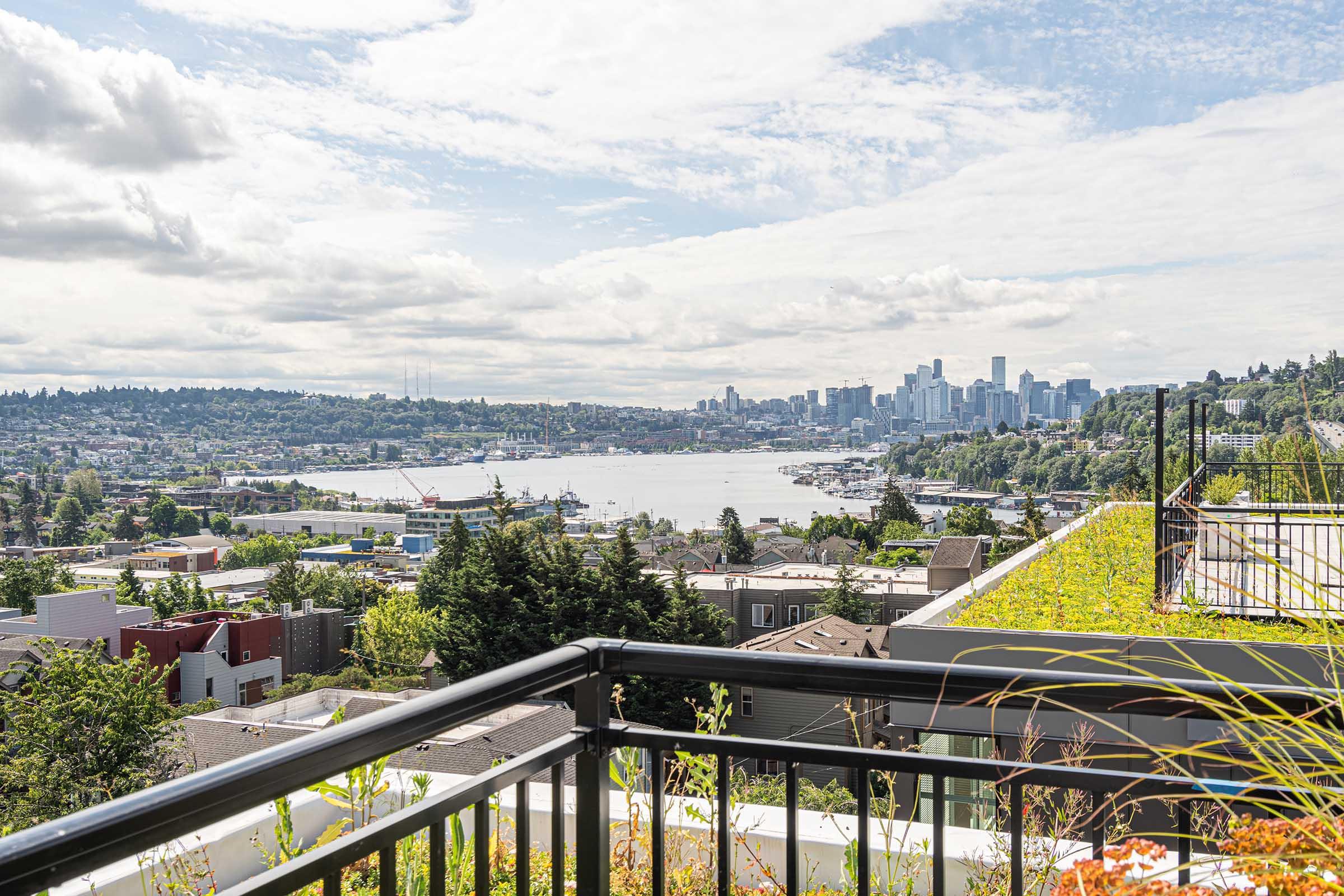 A panoramic view of a city skyline beside a body of water, with greenery and rooftops in the foreground. The scene captures a mix of urban and natural elements, featuring cloudy skies over Seattle's downtown, with boats visible on the water and lush trees in the landscape.