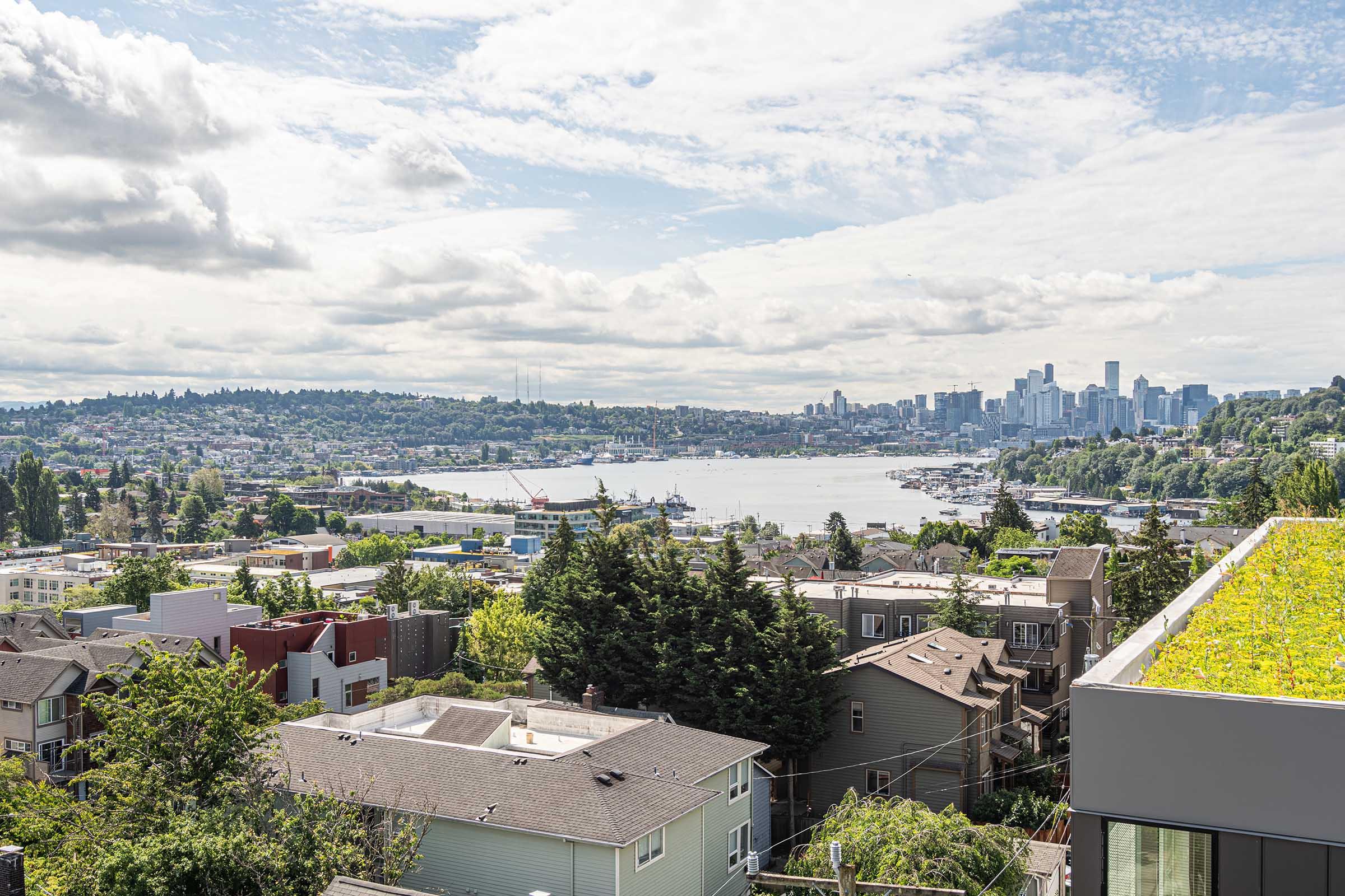 A panoramic view of Seattle featuring a picturesque waterfront, boats on the water, and the city skyline in the background. Lush green hills and residential buildings foreground the scene, with a partly cloudy sky overhead.