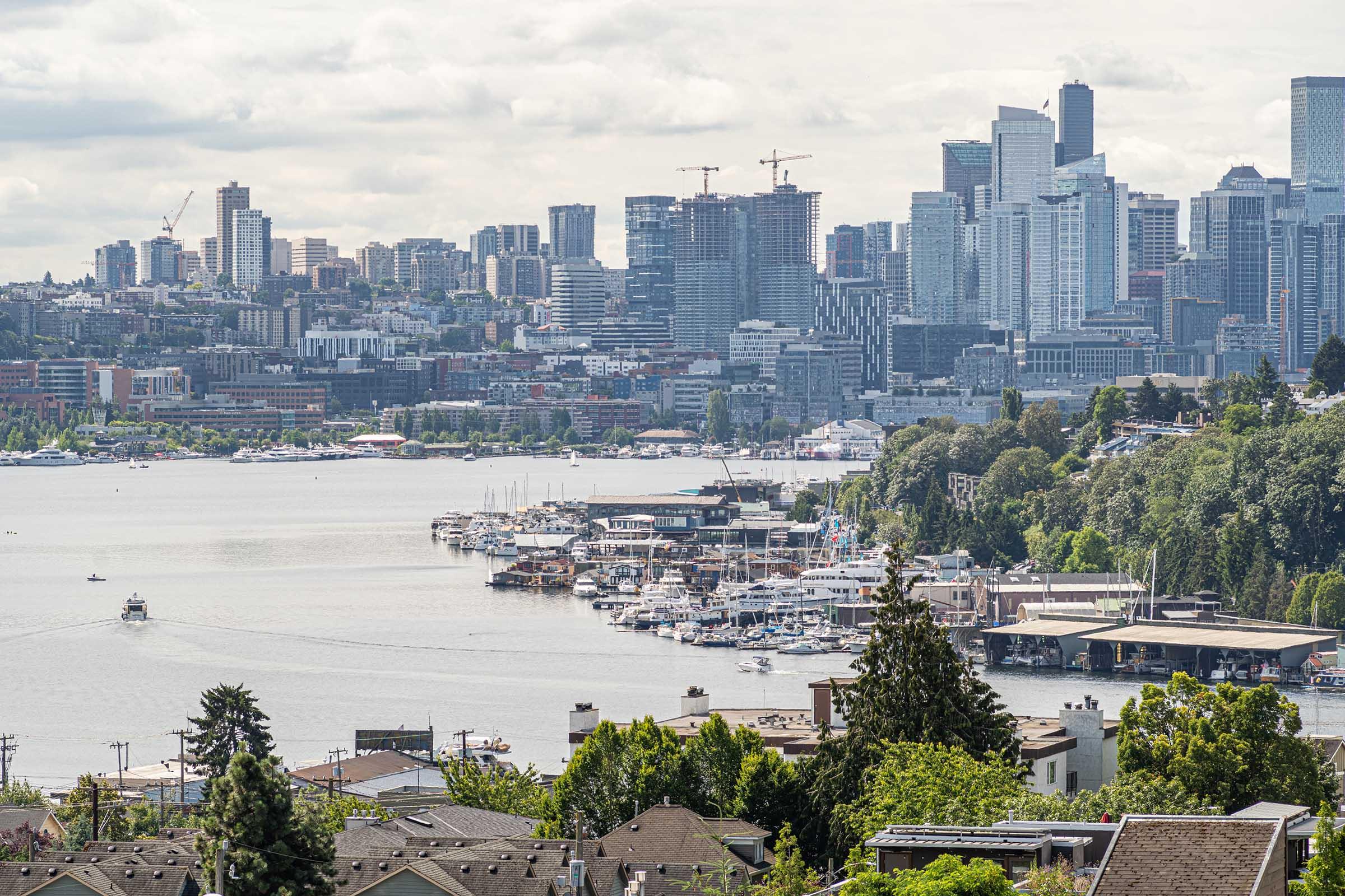 A panoramic view of a city skyline featuring modern skyscrapers, with a waterfront filled with boats and marinas. Lush trees and residential buildings are in the foreground, while cranes and construction sites hint at ongoing development. The scene is set under a partly cloudy sky.