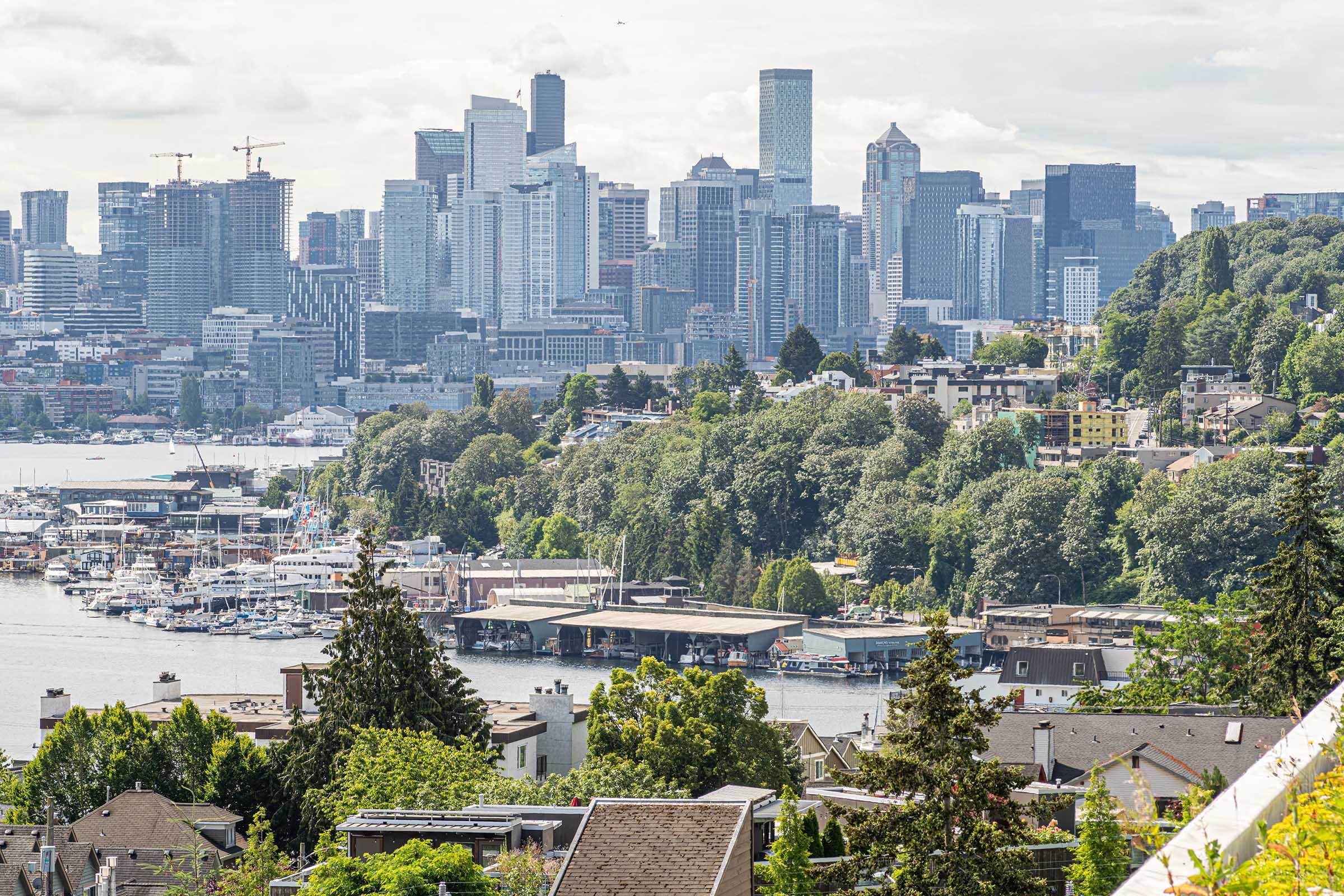 A panoramic view of Seattle's skyline featuring modern skyscrapers and waterfront. The foreground includes lush green trees and residential buildings, while the background showcases boats in the harbor and the distinct architecture of downtown Seattle under a partly cloudy sky.