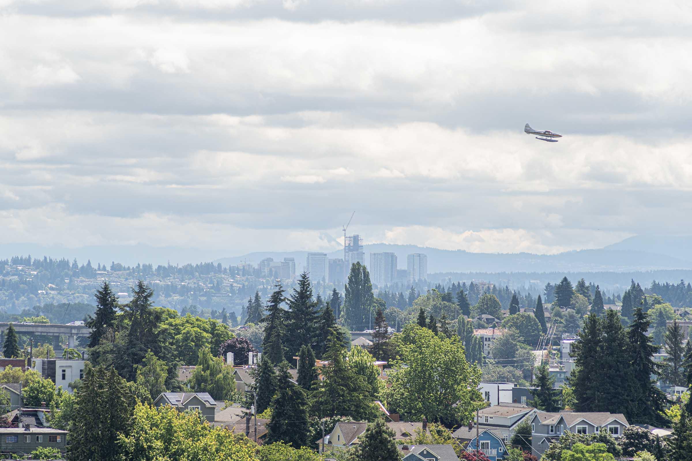 A scenic view of a city skyline with tall buildings in the background, surrounded by green trees and residential neighborhoods. A small airplane is flying in the sky above, with a cloudy atmosphere providing a moody backdrop to the landscape. Mountains are visible in the distance.