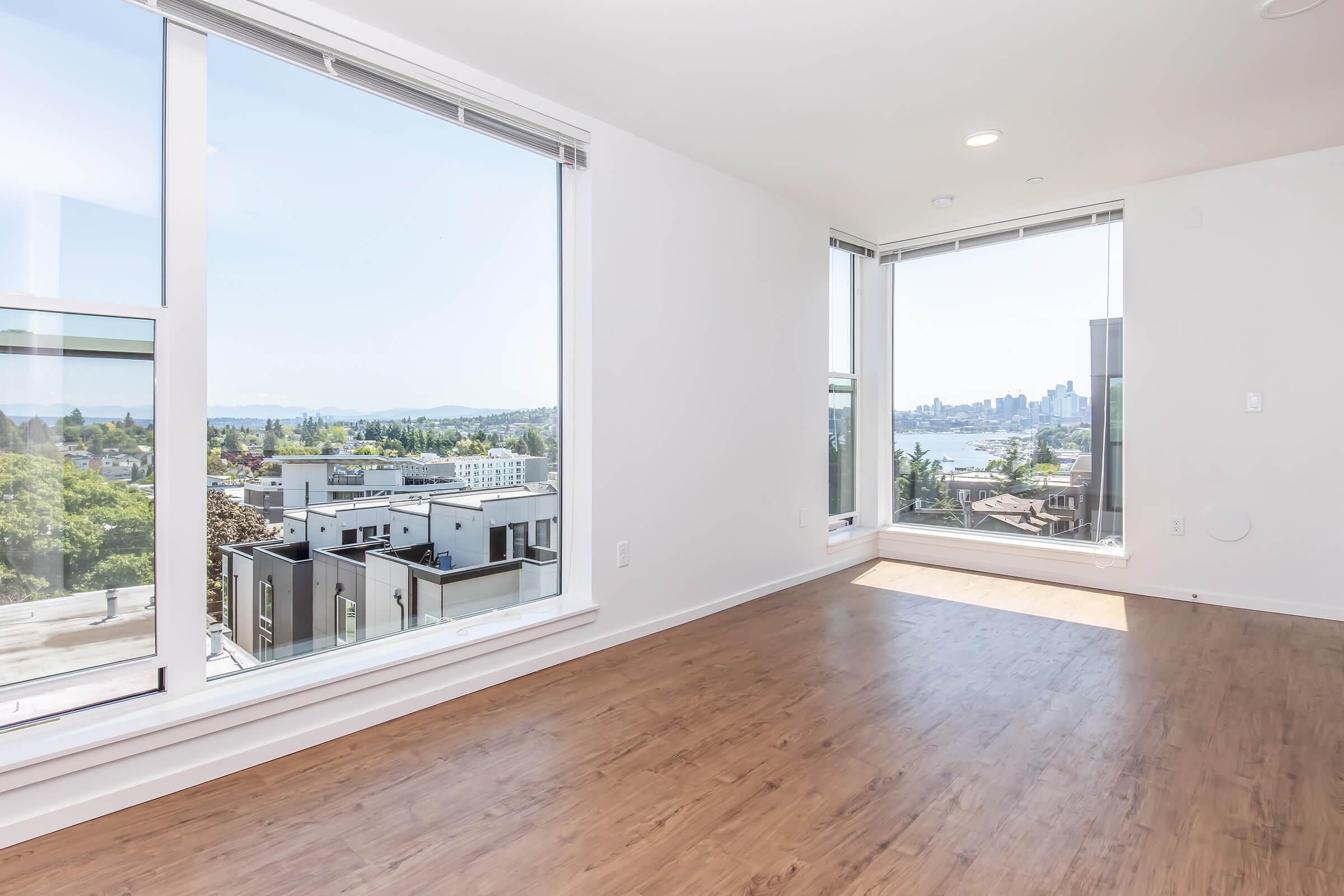 A bright, empty room with large windows offering a panoramic view of the city and water. The space features light-colored walls and a warm wood floor, creating a modern and inviting atmosphere. The natural light enhances the openness of the area.