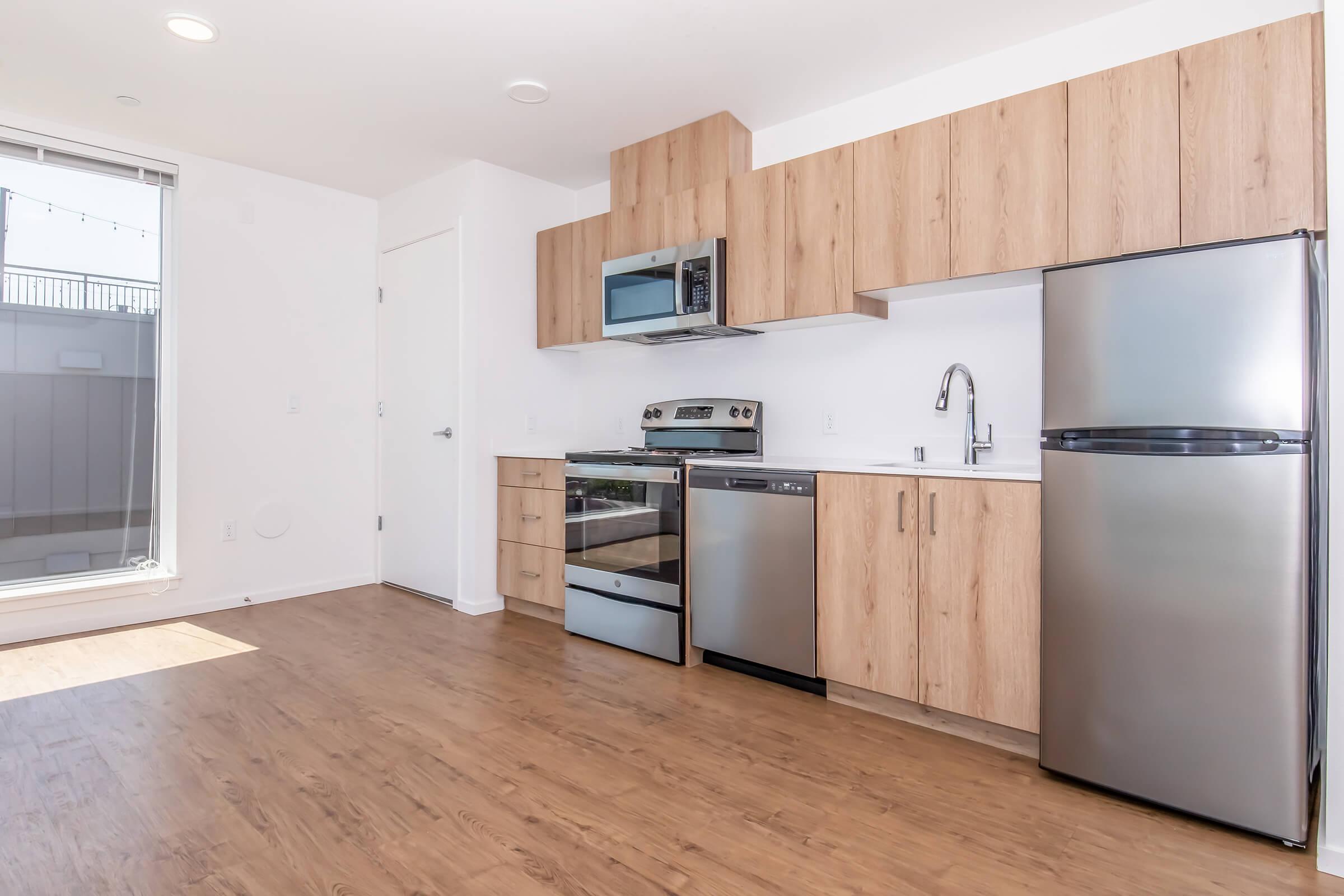 Modern kitchen featuring wooden cabinetry, stainless steel appliances including a microwave, oven, dishwasher, and refrigerator. The space has a light, airy feel with large windows allowing natural light, and a wood-like floor for a contemporary aesthetic.