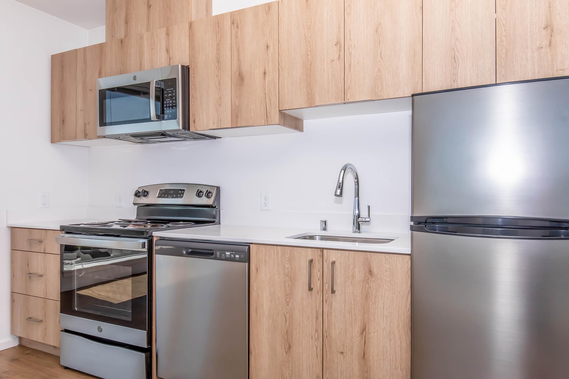 Modern kitchen featuring light wood cabinetry, stainless steel appliances including a microwave, oven, dishwasher, and refrigerator, with a sleek countertop and a single basin sink. The design is clean and contemporary, emphasizing a minimalist aesthetic.