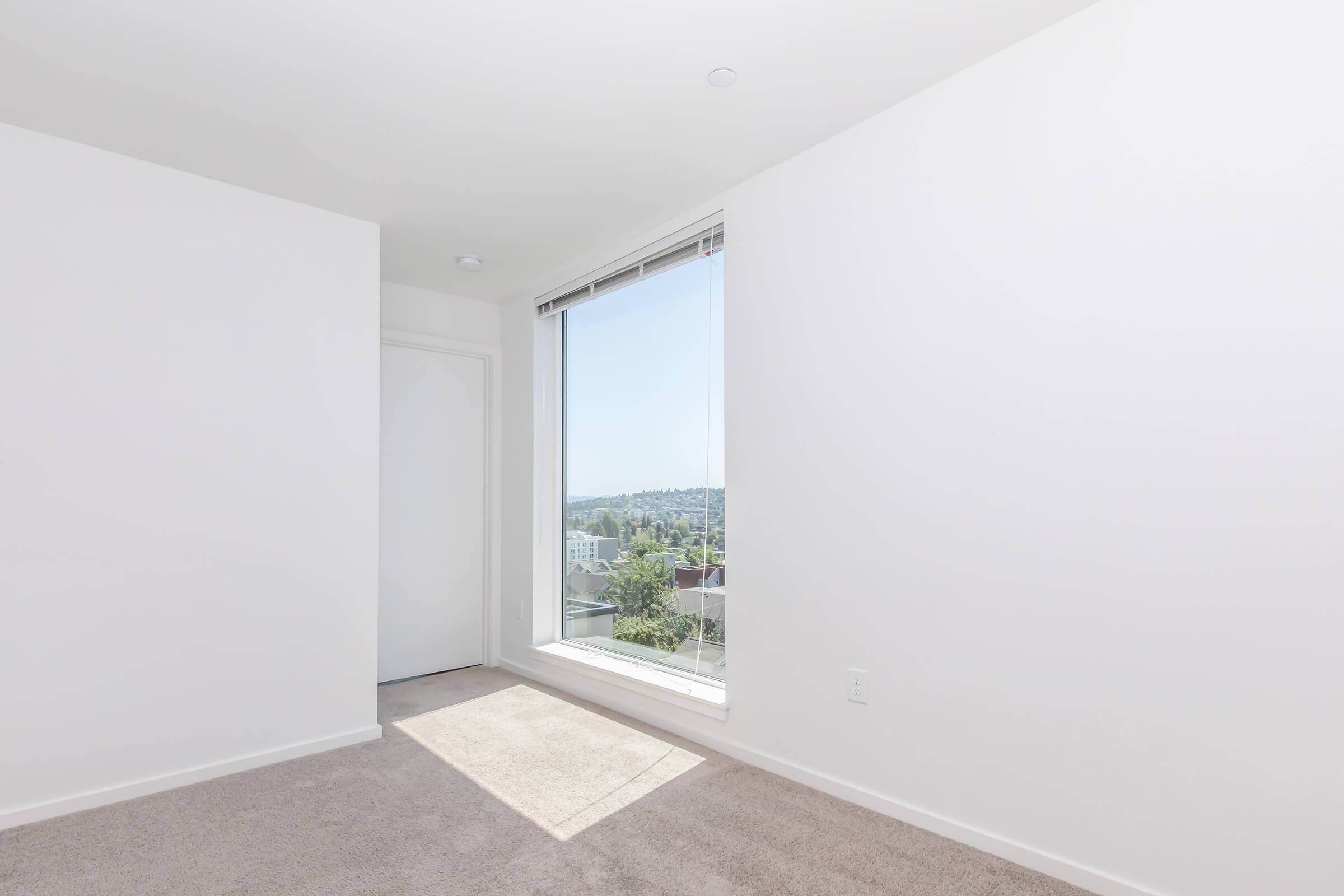 A bright, minimalistic room with white walls and light carpet. A large window provides natural light and offers a view of greenery outside. The space appears empty and modern, with simple design elements and no furniture visible.