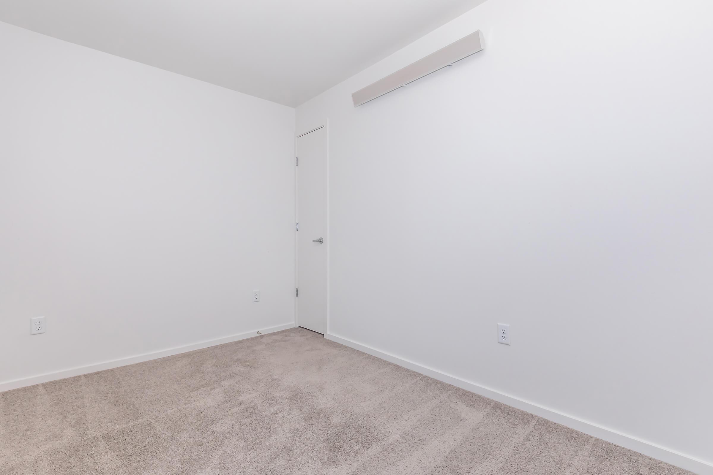 Empty room with light beige carpet and plain white walls. A door is visible on the left side, and there is a light fixture along the upper wall. The overall space appears minimalistic and uncluttered.