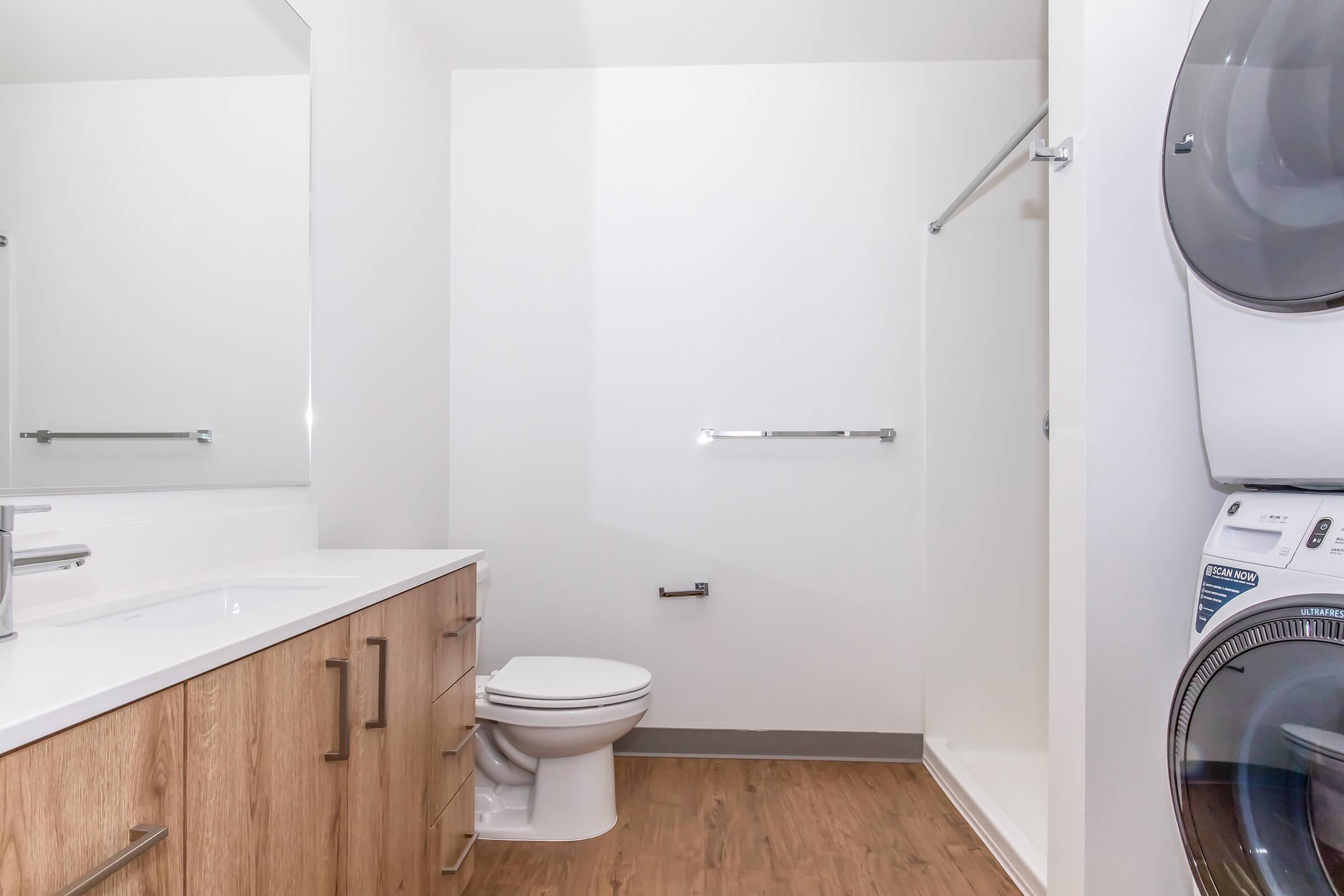 A modern, minimalist bathroom featuring a toilet, a sink with wooden cabinetry, a large mirror, and a shower without a curtain. A washer and dryer are stacked against one wall. The space has light-colored walls and a wood-like floor, creating a clean and airy atmosphere.