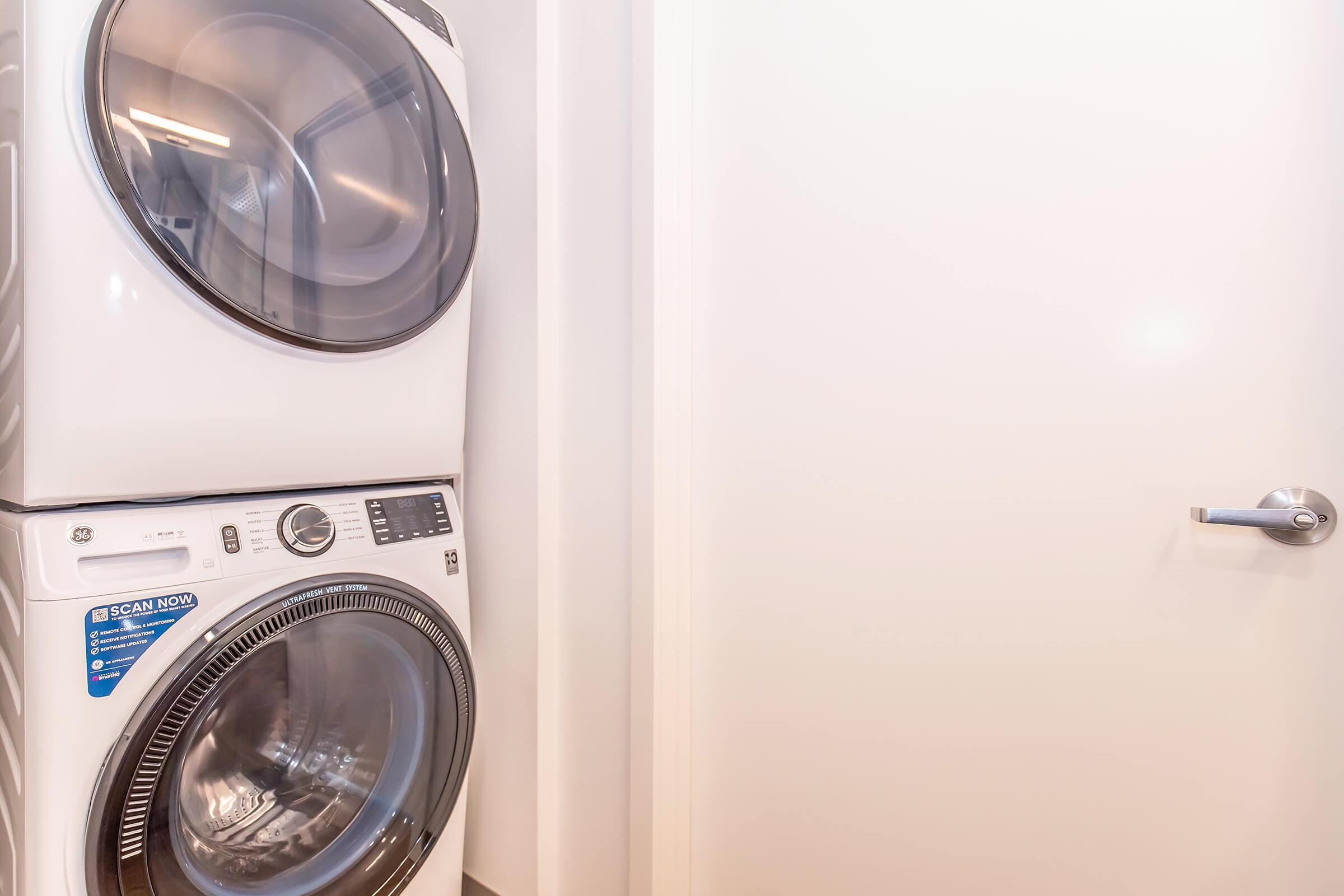 A modern white washer and dryer stacked together in a bright laundry area. The door leading out of the room is partially visible, with a sleek handle. The overall setting is clean and minimalistic, emphasizing the appliances.