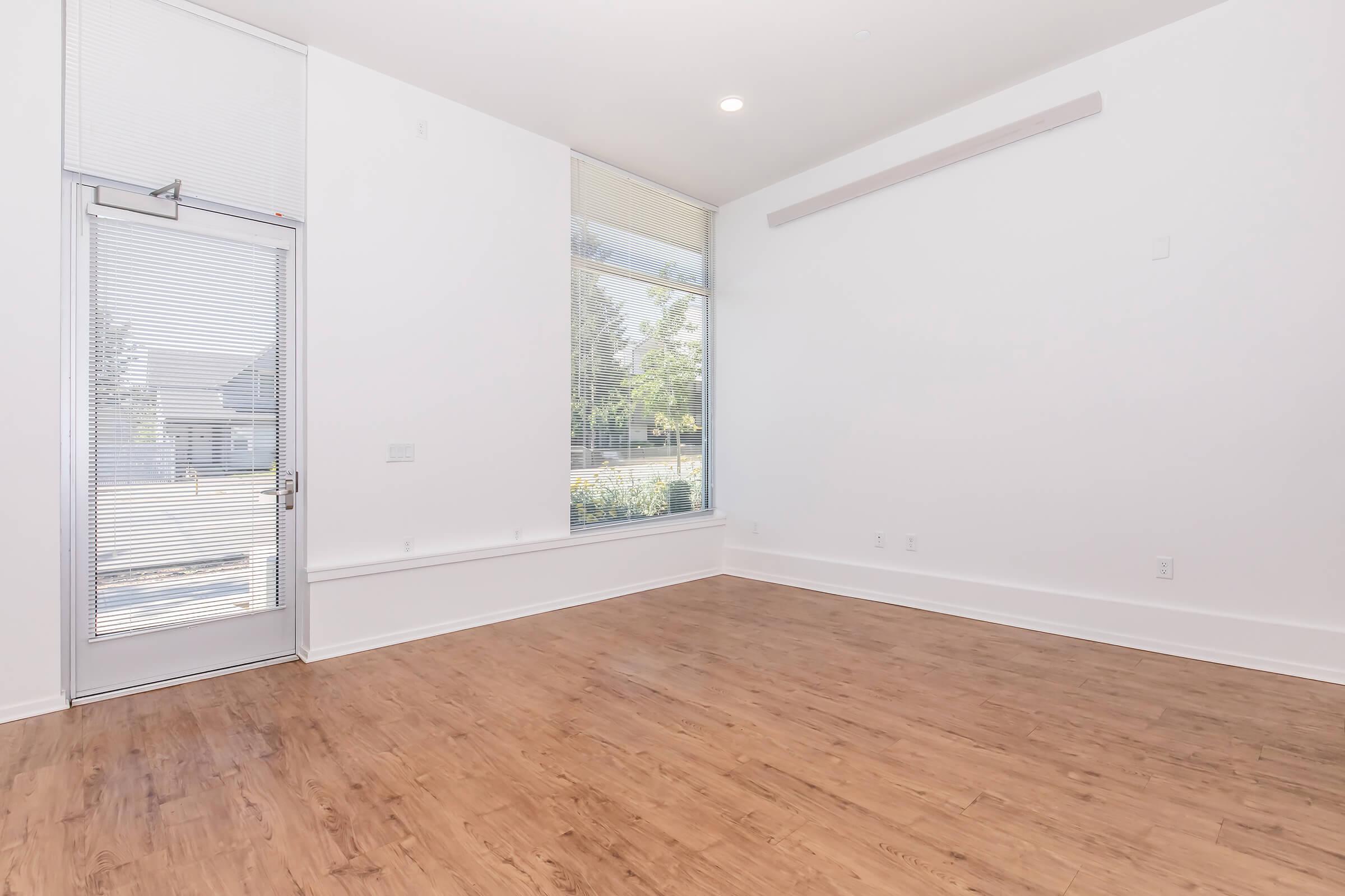 A bright, empty room with large windows allowing natural light, light-colored walls, and a wood floor. A glass door is visible on one side, leading outside, and there is minimal furniture or decoration, creating a spacious and airy feel.