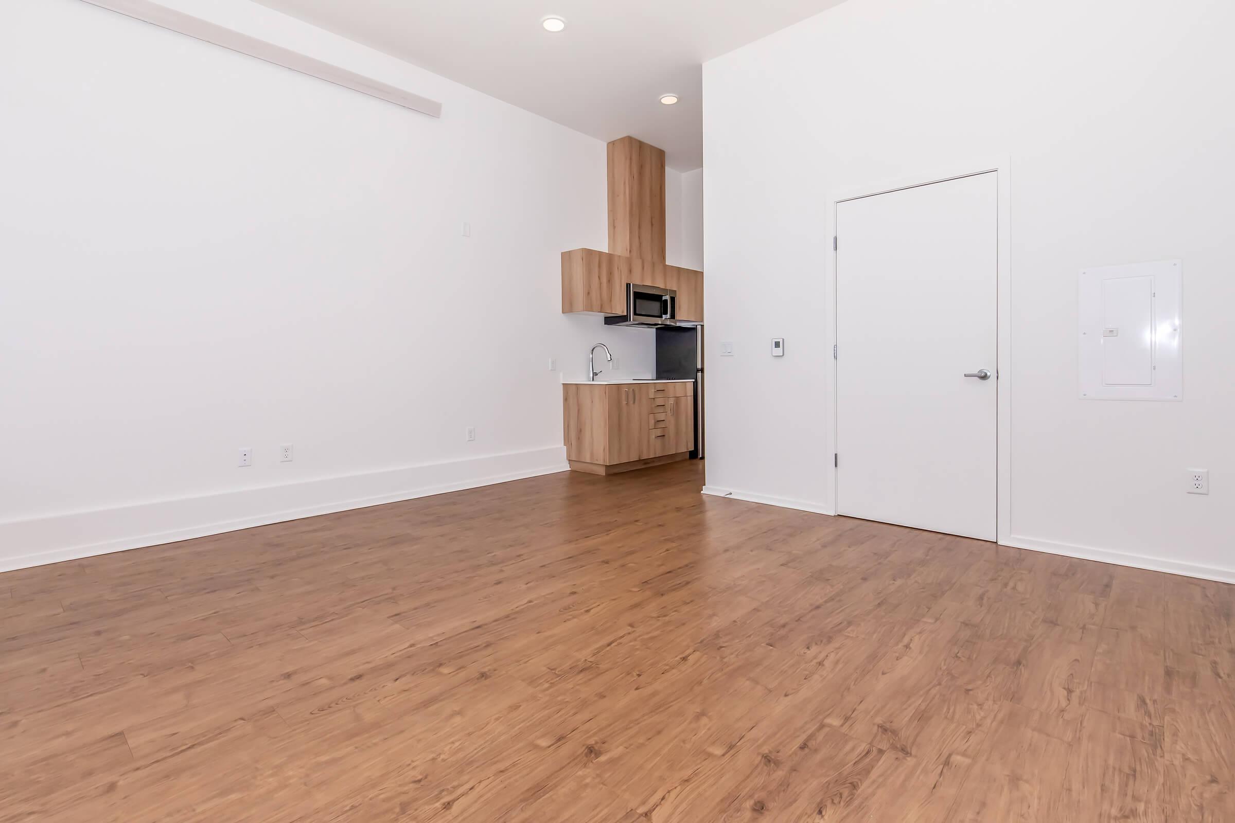 An empty modern apartment interior with light hardwood flooring, white walls, and minimalistic design. The kitchen area features wooden cabinets and a stainless steel microwave. A closed white door is visible, along with a wall panel. The space is well-lit with overhead lights.