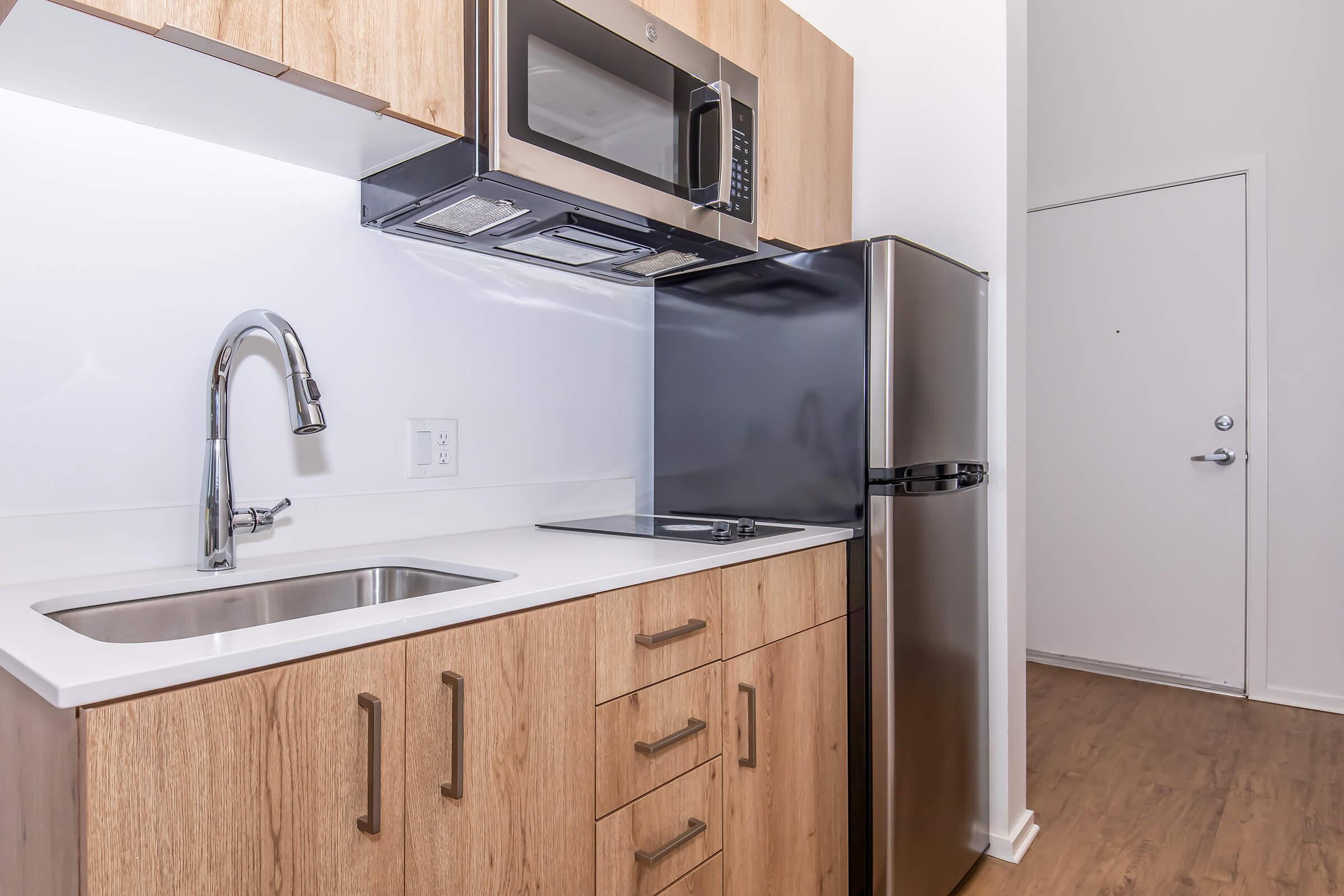 Modern kitchen featuring a stainless steel refrigerator, microwave, and stovetop. Light wood cabinetry provides storage, while a sleek sink is positioned under the countertop. Bright and minimalistic design with a clean aesthetic, perfect for compact living spaces.