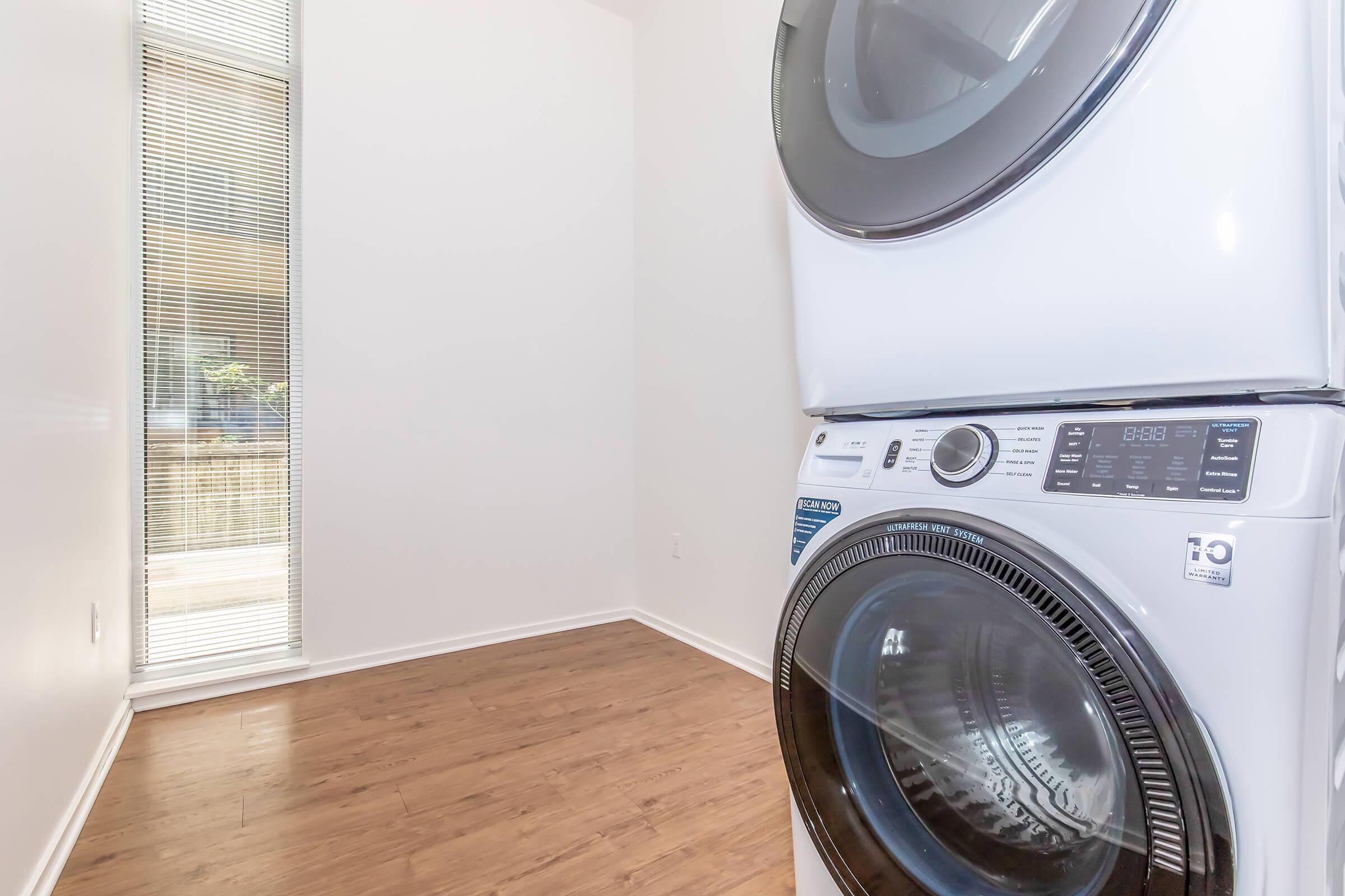 A modern laundry room featuring a stacked washer and dryer in white, positioned against a light-colored wall. A window with vertical blinds allows natural light into the space, while the floor is made of wood laminate, enhancing the contemporary feel.
