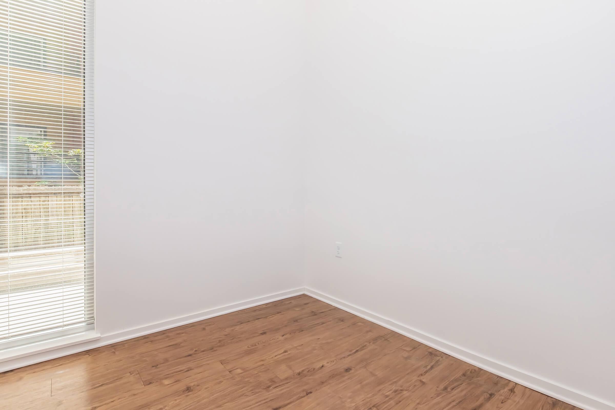 A bright, empty corner of a room with light-colored walls and a wooden floor. A window with blinds allows natural light to enter, but the view outside is not visible. The space appears minimalistic and open, ideal for various purposes such as a reading nook or small office.