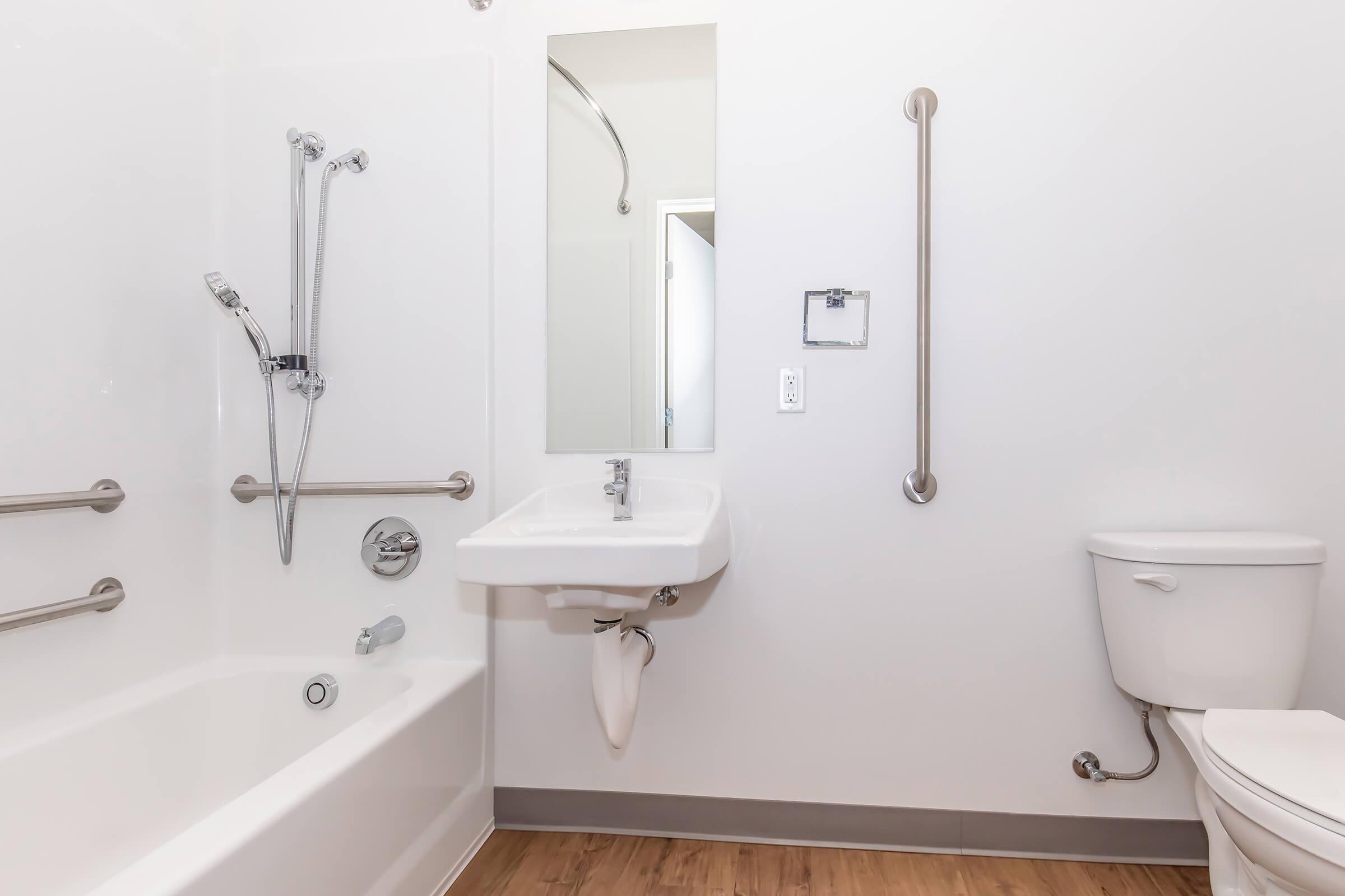 A clean and modern bathroom featuring a white bathtub with grab bars, a wall-mounted sink, a toilet, and a large mirror. The space has light-colored walls and wooden flooring, emphasizing a fresh and spacious atmosphere.