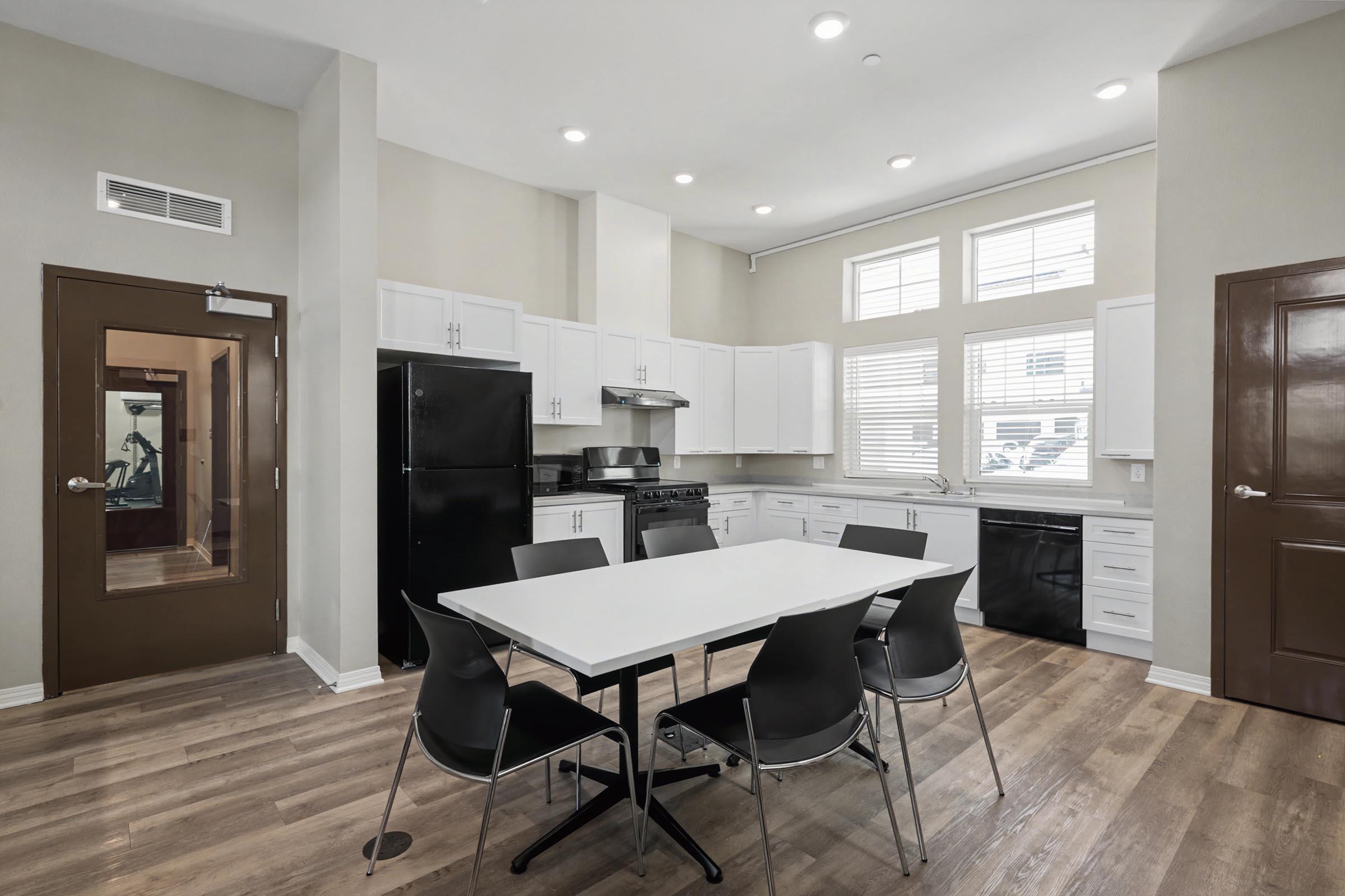 Modern kitchen and dining area featuring a white table with black chairs, black refrigerator, dishwasher, and stove. The space has large windows for natural light, light-colored walls, and wooden flooring, creating an open and inviting atmosphere. A door leads to a separate room visible in the background.