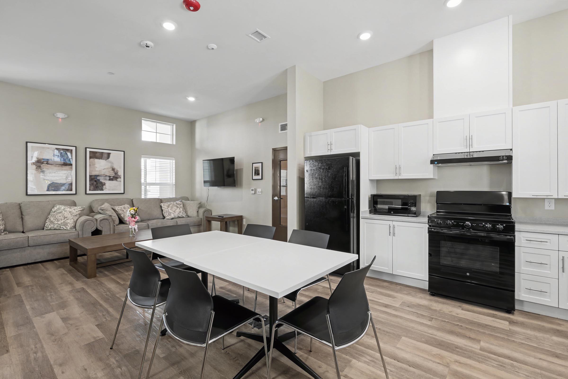 A modern kitchen and living area featuring a white dining table with six black chairs, a gray sofa, a coffee table, and a refrigerator. The kitchen includes white cabinets, a black stove, and a microwave. Large windows let in natural light, creating a bright and inviting atmosphere.
