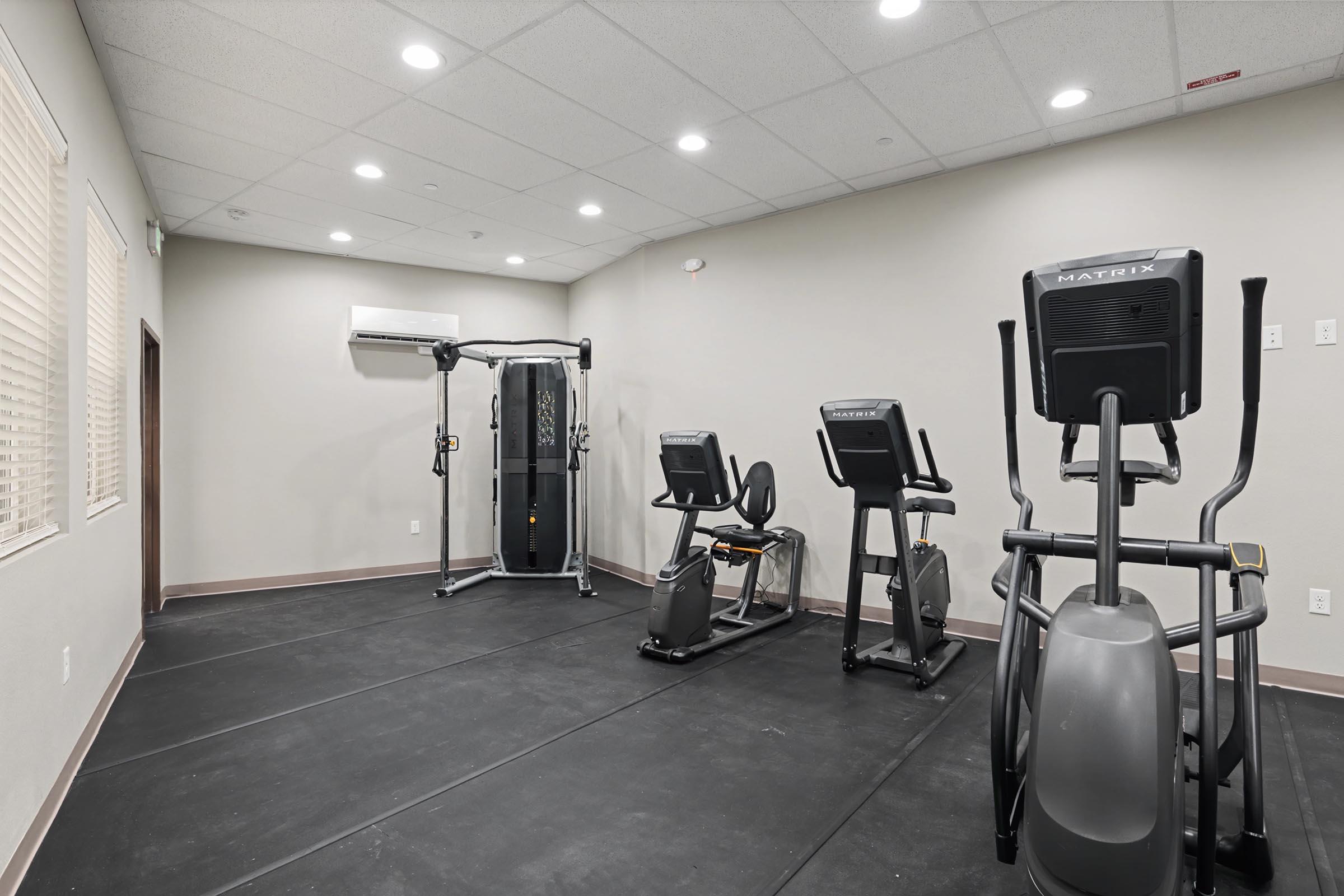 A modern gym interior featuring exercise equipment. There is a cable machine, an elliptical trainer, and a stationary bike arranged on a black rubber flooring. The walls are light-colored with a few windows allowing natural light, and the ceiling is well-lit with recessed lighting.