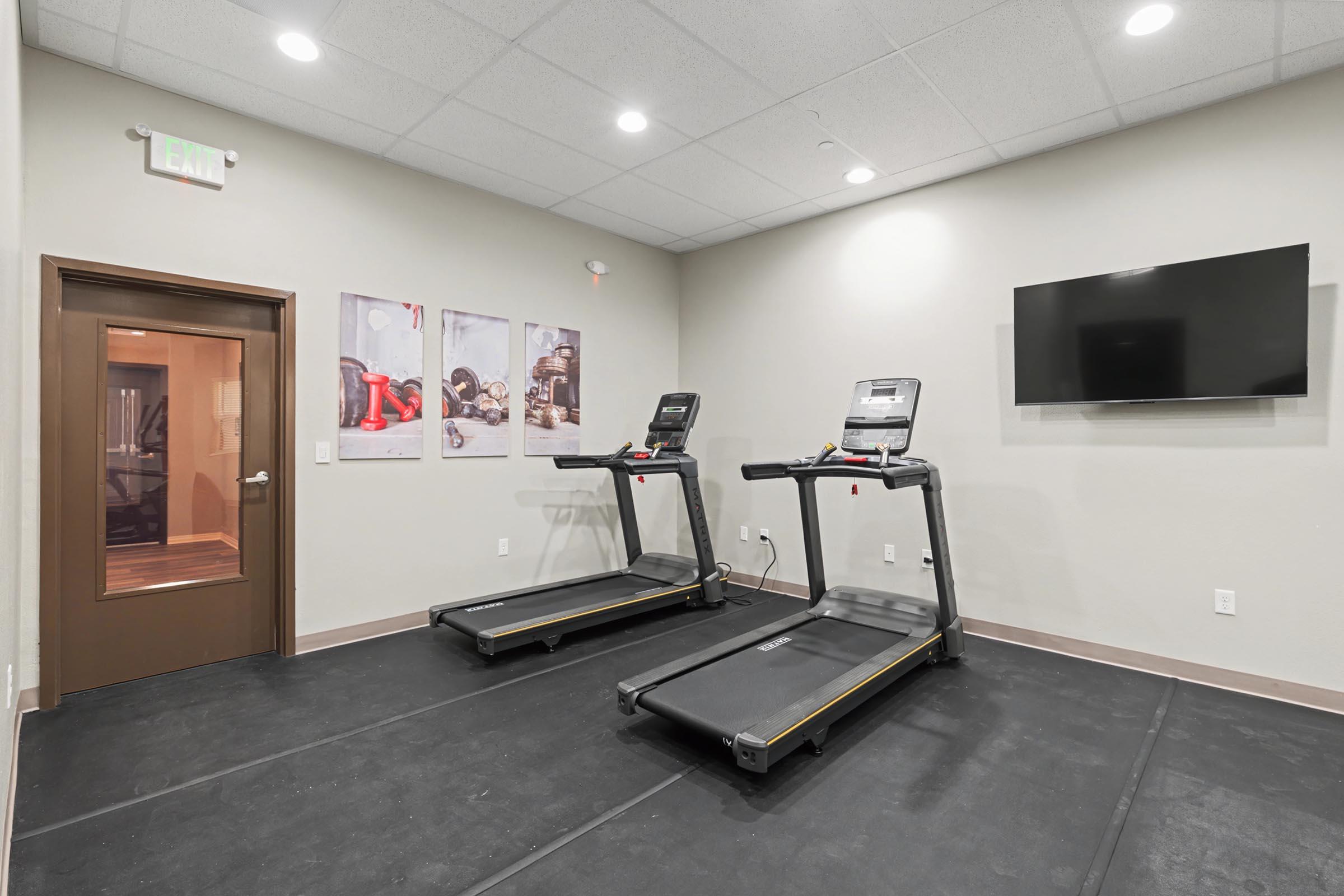 A modern fitness room featuring two treadmills positioned on a black rubber floor. A wall-mounted television is visible, and there are fitness-themed images displayed on the wall. A door leads to another area, possibly a sauna or changing room. The space is well-lit with neutral-colored walls.