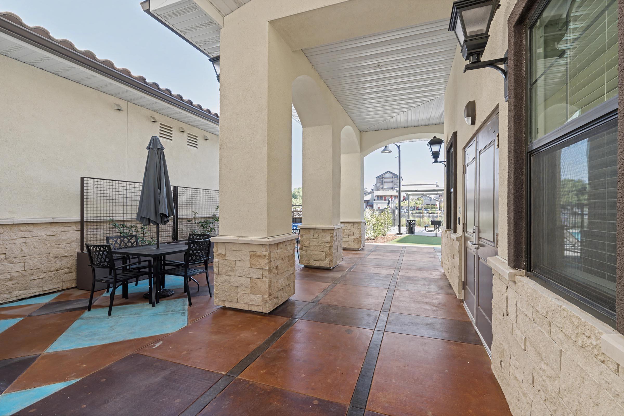 A shaded outdoor area features a table with four chairs and a large umbrella, situated next to a light-colored building. The walkway has decorative tiles and features lamps along the sides. In the background, there are hints of greenery and other structures visible.