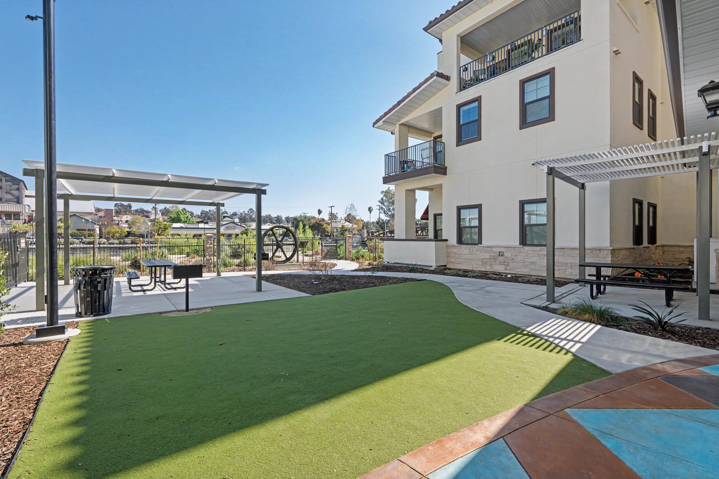 A landscaped outdoor area featuring artificial turf, picnic tables, and shaded seating structures. In the background, a modern multi-story building is visible, along with trees and open spaces. The scene is set under a clear blue sky, creating a welcoming atmosphere.