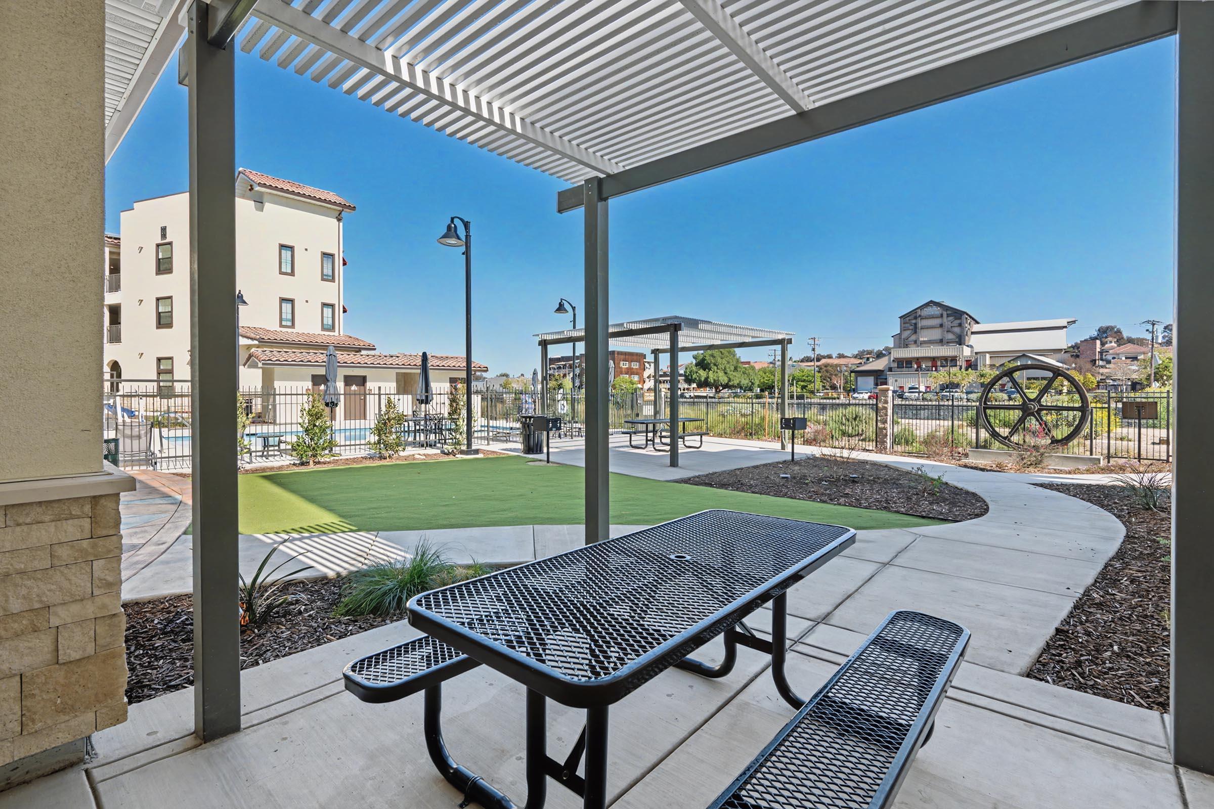 Outdoor seating area with a black picnic table and benches under a shaded structure. In the background, there is a grassy area with landscaped plants and a small gazebo. Nearby, a decorative piece resembling a large wheel adds interest to the space, which is situated near a modern residential building. Clear blue sky overhead.