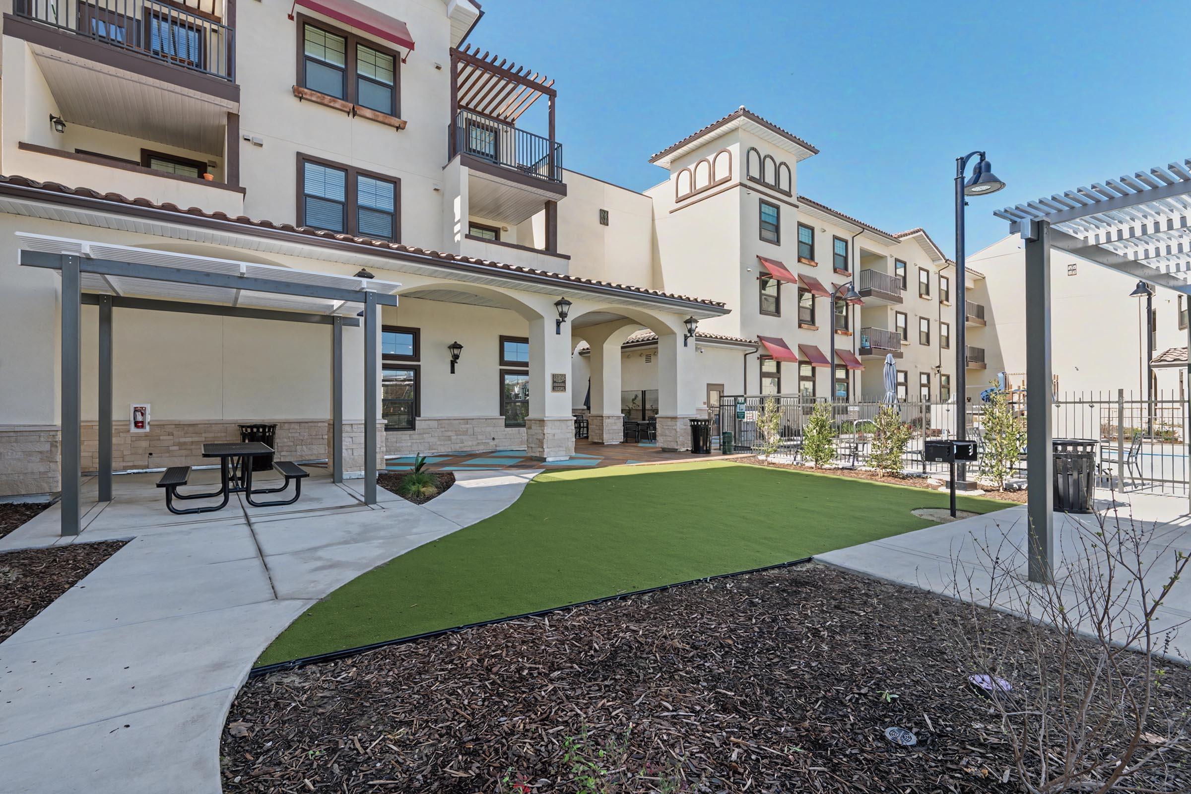 A landscaped outdoor area featuring a green lawn, picnic table, and shaded seating near a residential building. The space includes a pathway, decorative plants, and a swimming pool visible in the background, all under a clear blue sky.