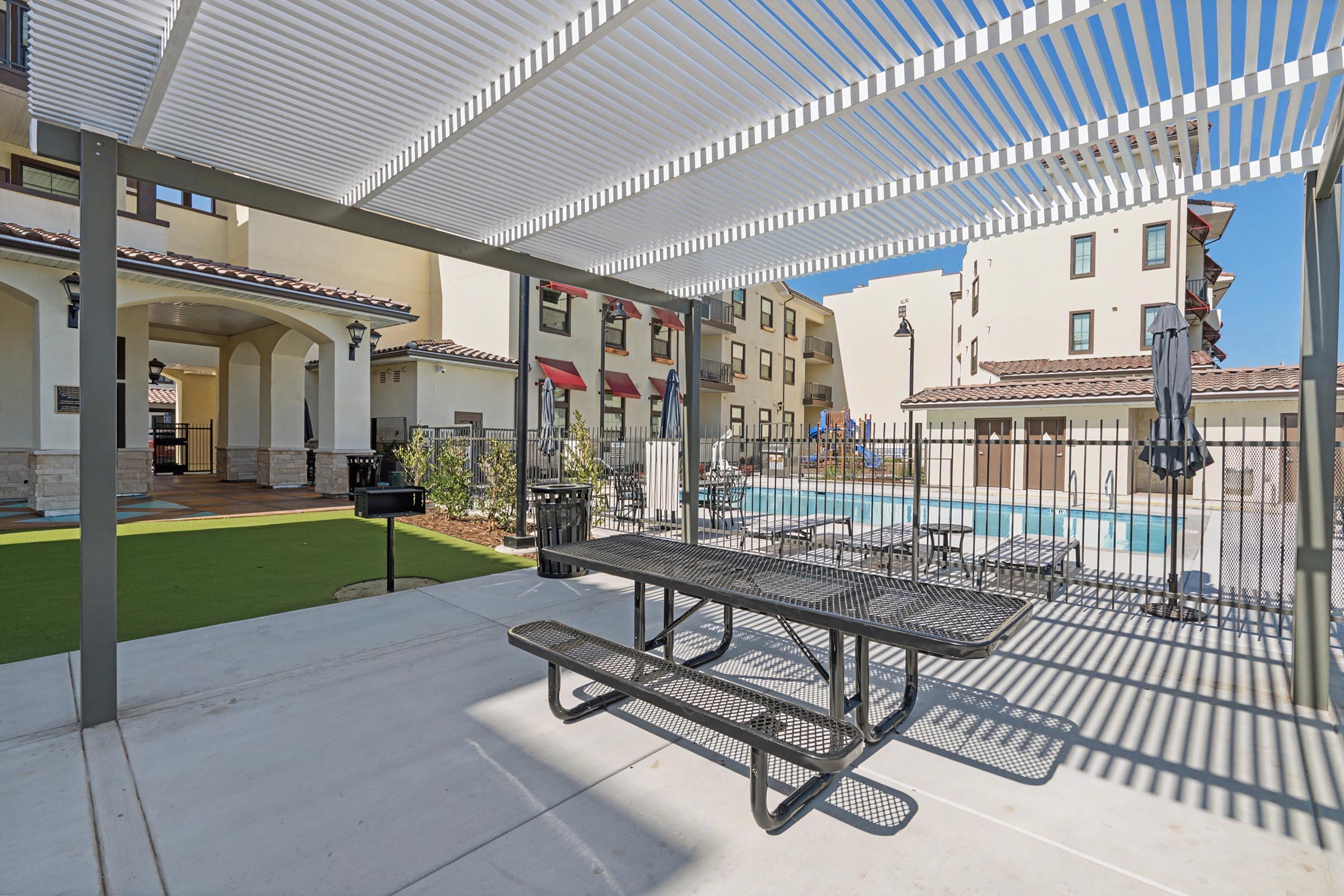 A shaded outdoor area with a picnic table and grill next to a swimming pool. The background features modern apartment buildings, with an arrangement of lounge chairs visible by the pool. Clear blue skies and bright sunlight enhance the inviting atmosphere of the space.