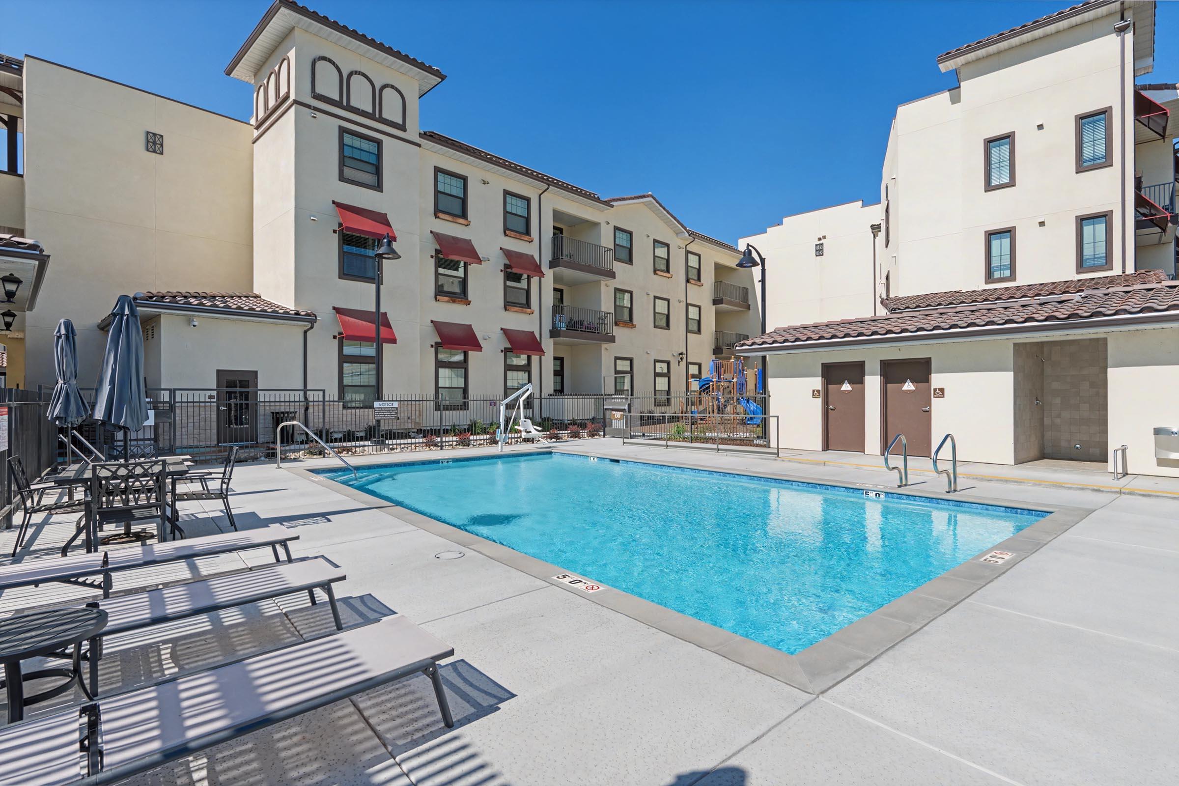 A bright, sunny pool area with a clear blue pool surrounded by lounge chairs. In the background are two multi-story buildings with balconies and red awnings, along with a playground nearby. The sky is clear and blue, creating a relaxing outdoor atmosphere.