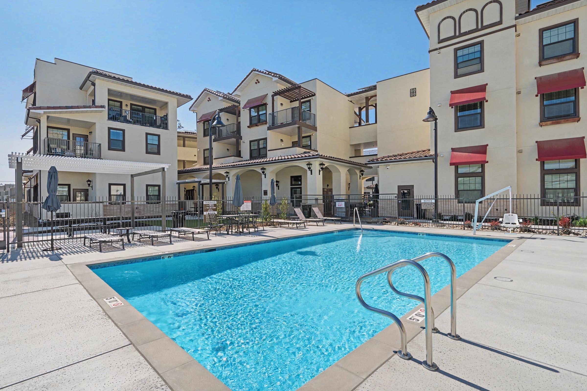 A residential complex featuring a swimming pool surrounded by lounge chairs and an outdoor area. The building includes multiple stories with balconies and large windows, offering a sunny and inviting atmosphere. Bright blue sky overhead enhances the relaxing vibe of the pool area.