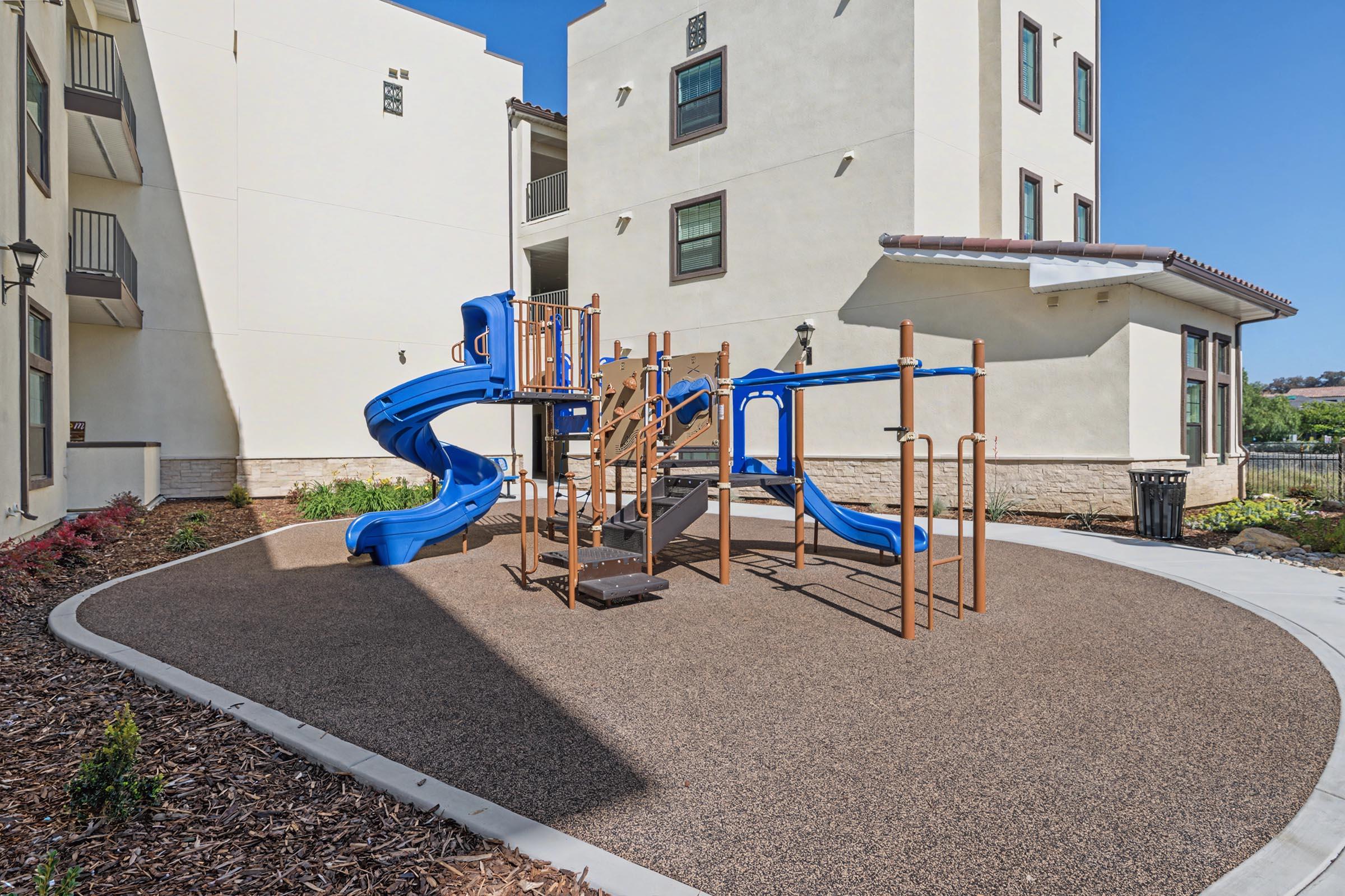 Playground area featuring a blue slide and climbing structure on a rubberized surface, surrounded by beige buildings. Well-maintained landscaping with shrubs in the foreground and clear blue sky above. Ideal for children's play and recreational activities.