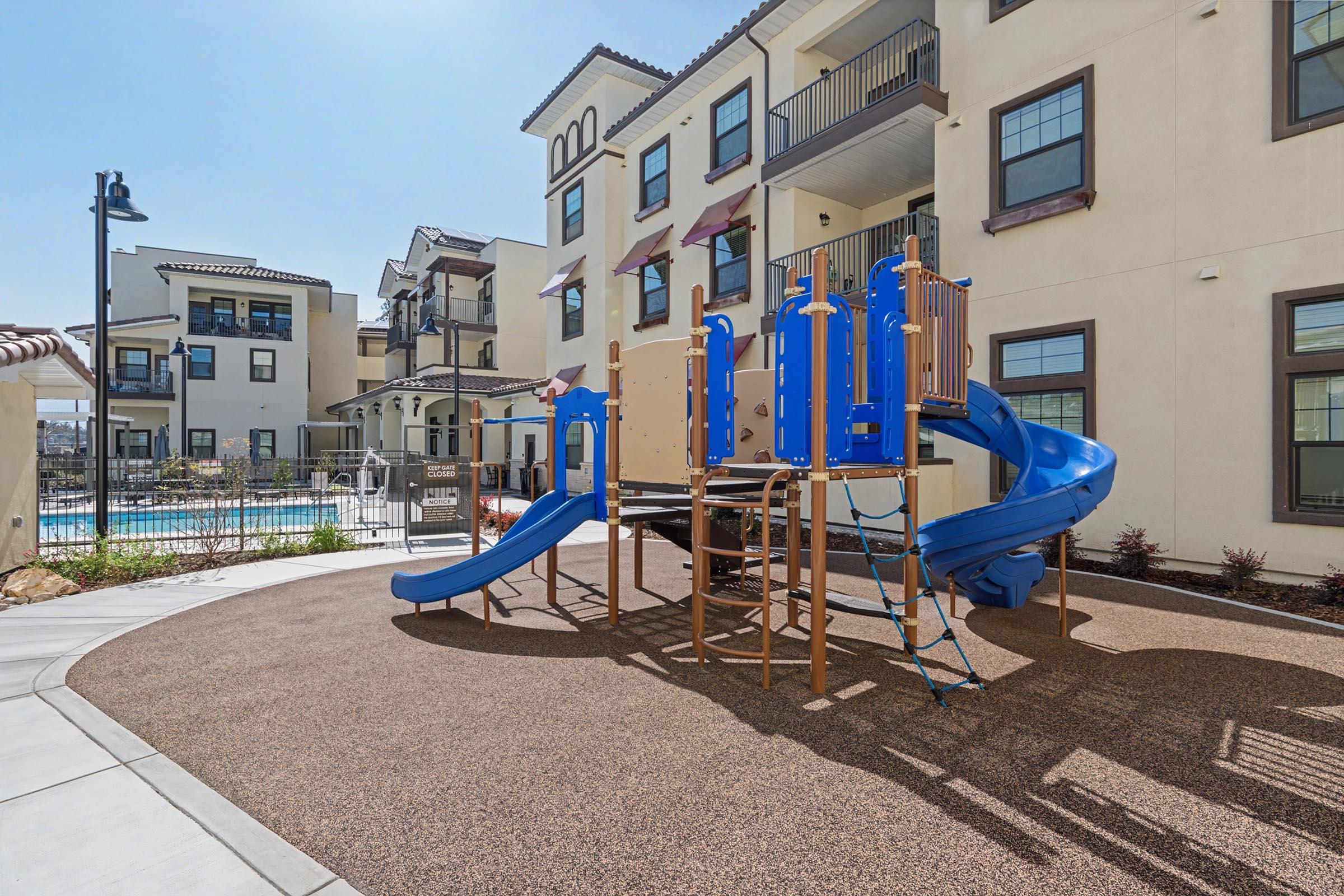 Playground area featuring a blue slide and climbing structure surrounded by sandy ground, adjacent to modern apartment buildings. A swimming pool is visible in the background, and the scene is illuminated by bright sunlight, creating a cheerful atmosphere for outdoor play.