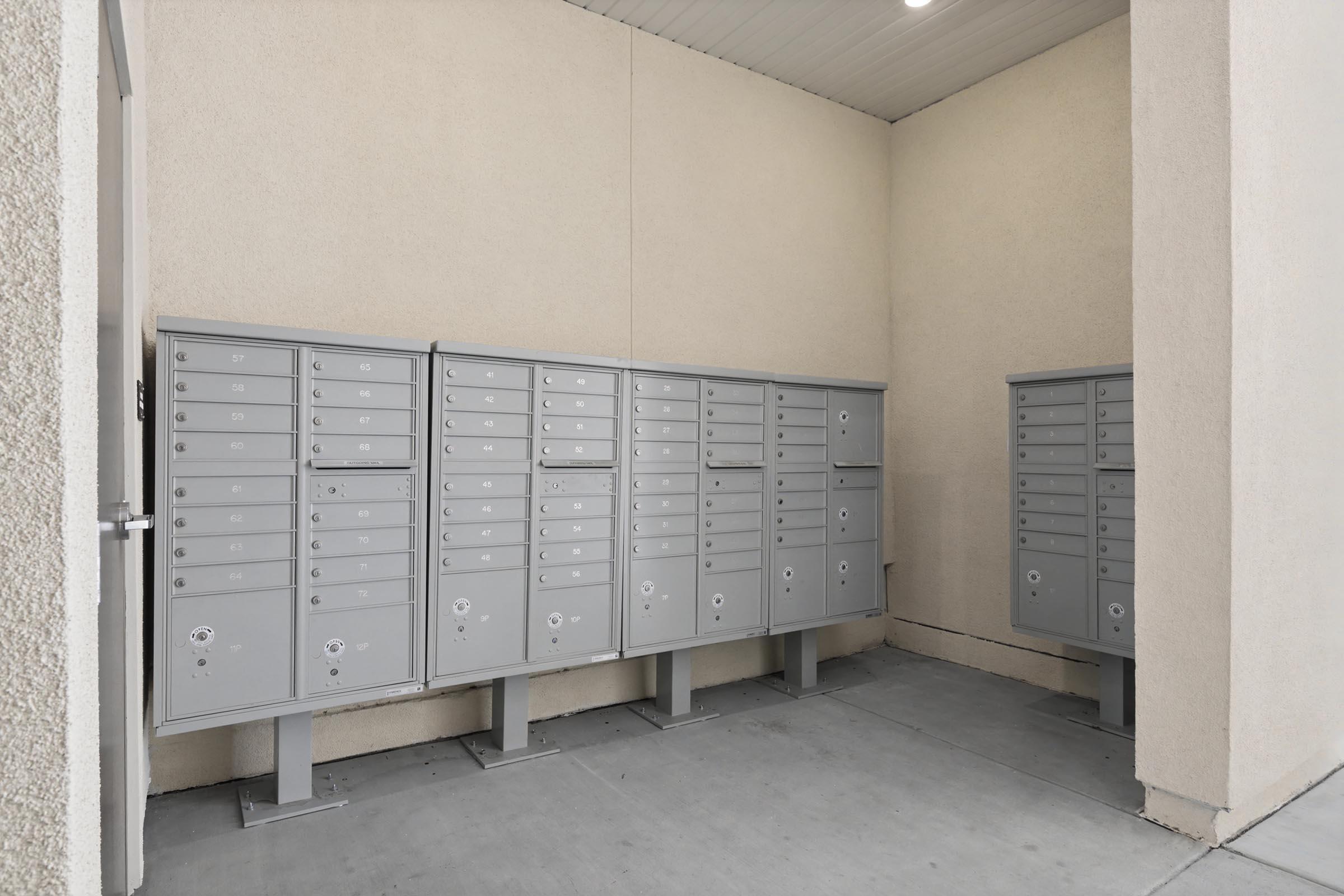 A row of gray, wall-mounted mailboxes in a corner of a building. The mailboxes are arranged in two groups, secured with locks, and feature multiple slots for letters and packages. The area has a plain, light-colored wall and a concrete floor, creating a simple, functional aesthetic.
