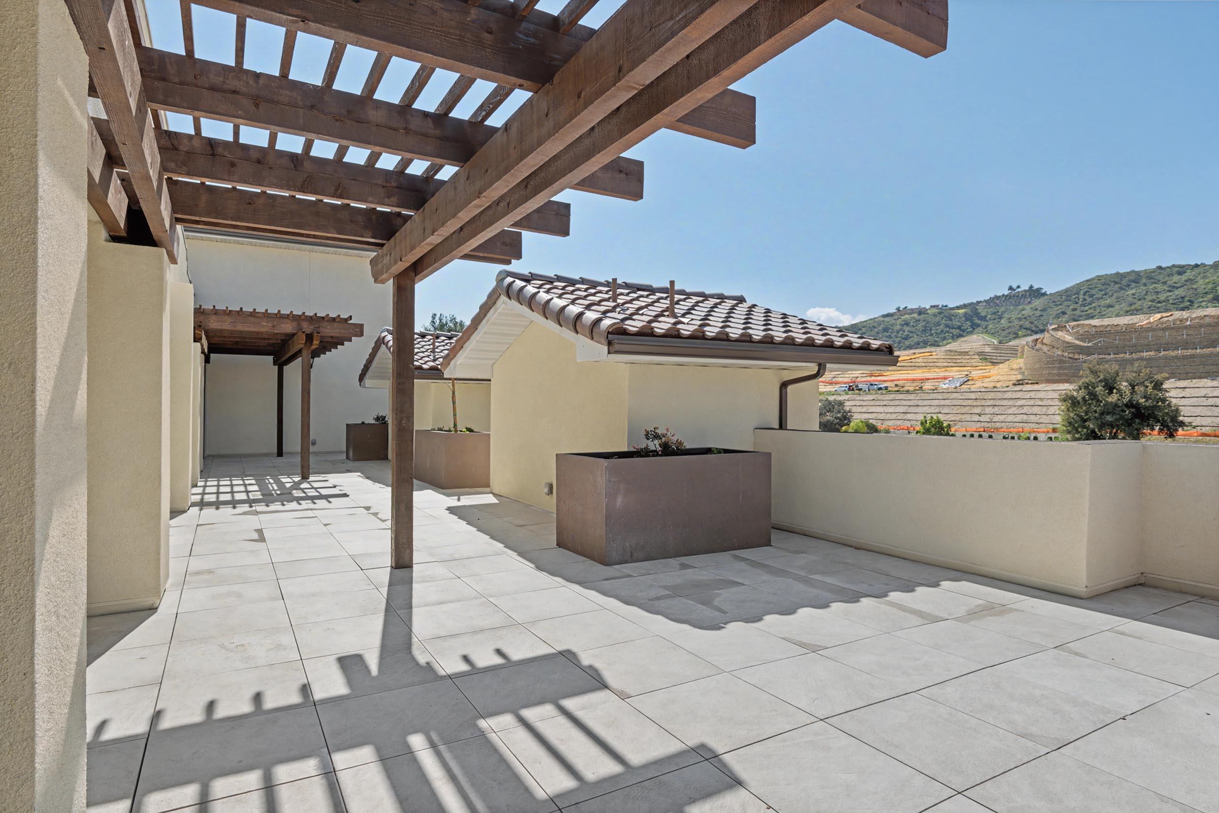 A spacious outdoor terrace with a pergola, featuring tiled flooring and planters. The backdrop includes rolling hills and a clear blue sky, creating a bright and inviting atmosphere. Sunlight casts shadows on the floor, enhancing the tranquil setting.