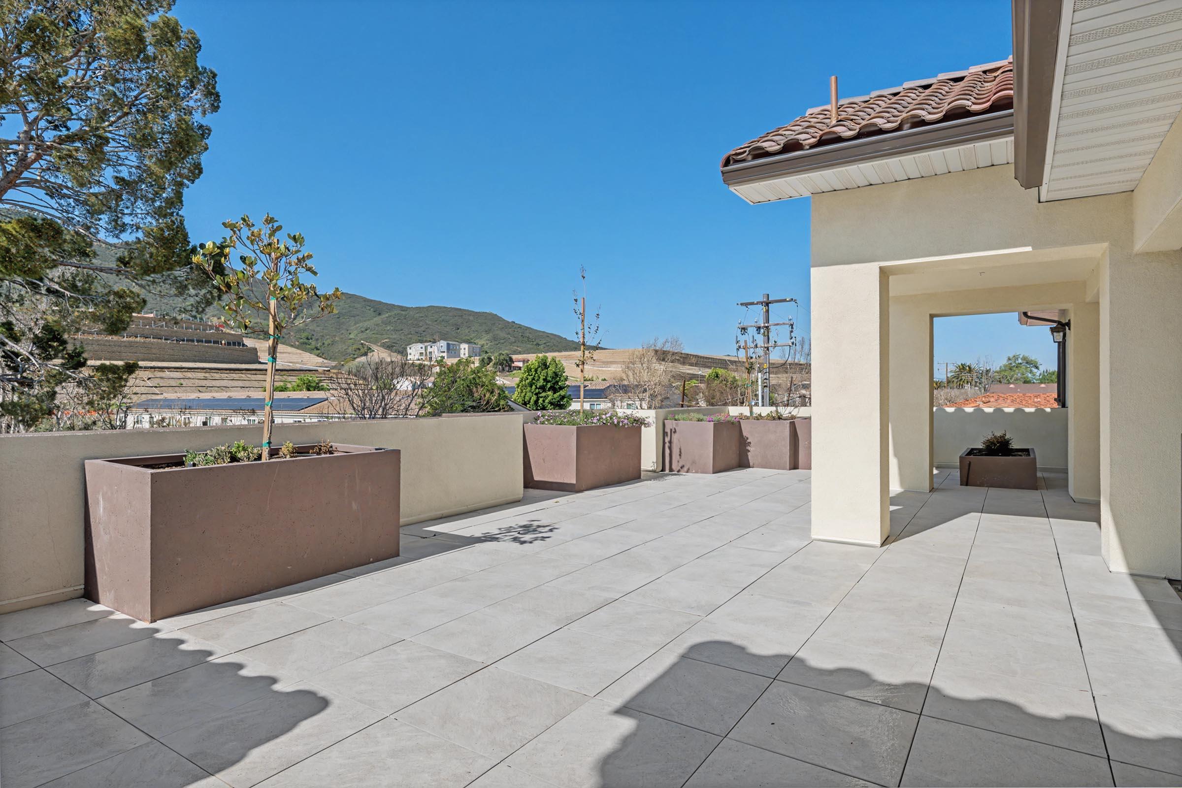 A spacious outdoor terrace featuring large planters, tiled flooring, and a clear blue sky. In the background, rolling hills and power lines are visible, providing a serene, natural setting. The terrace is bathed in sunlight, creating an inviting atmosphere.