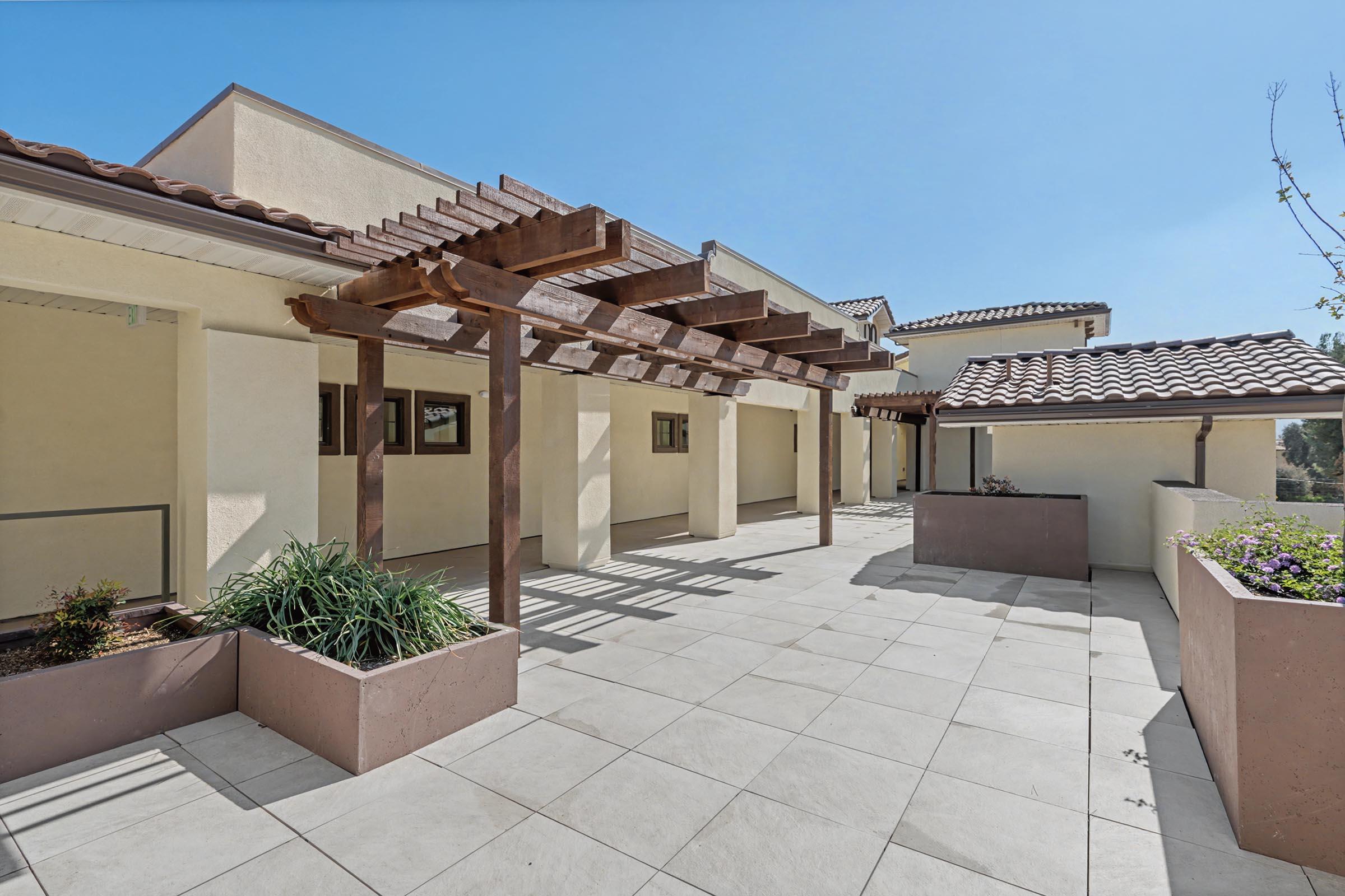 A sunny outdoor area featuring a spacious patio with tiled flooring, surrounded by modern architectural elements. There are wooden pergolas casting shadows, planters with greenery, and a clear blue sky overhead, creating a bright and inviting atmosphere.